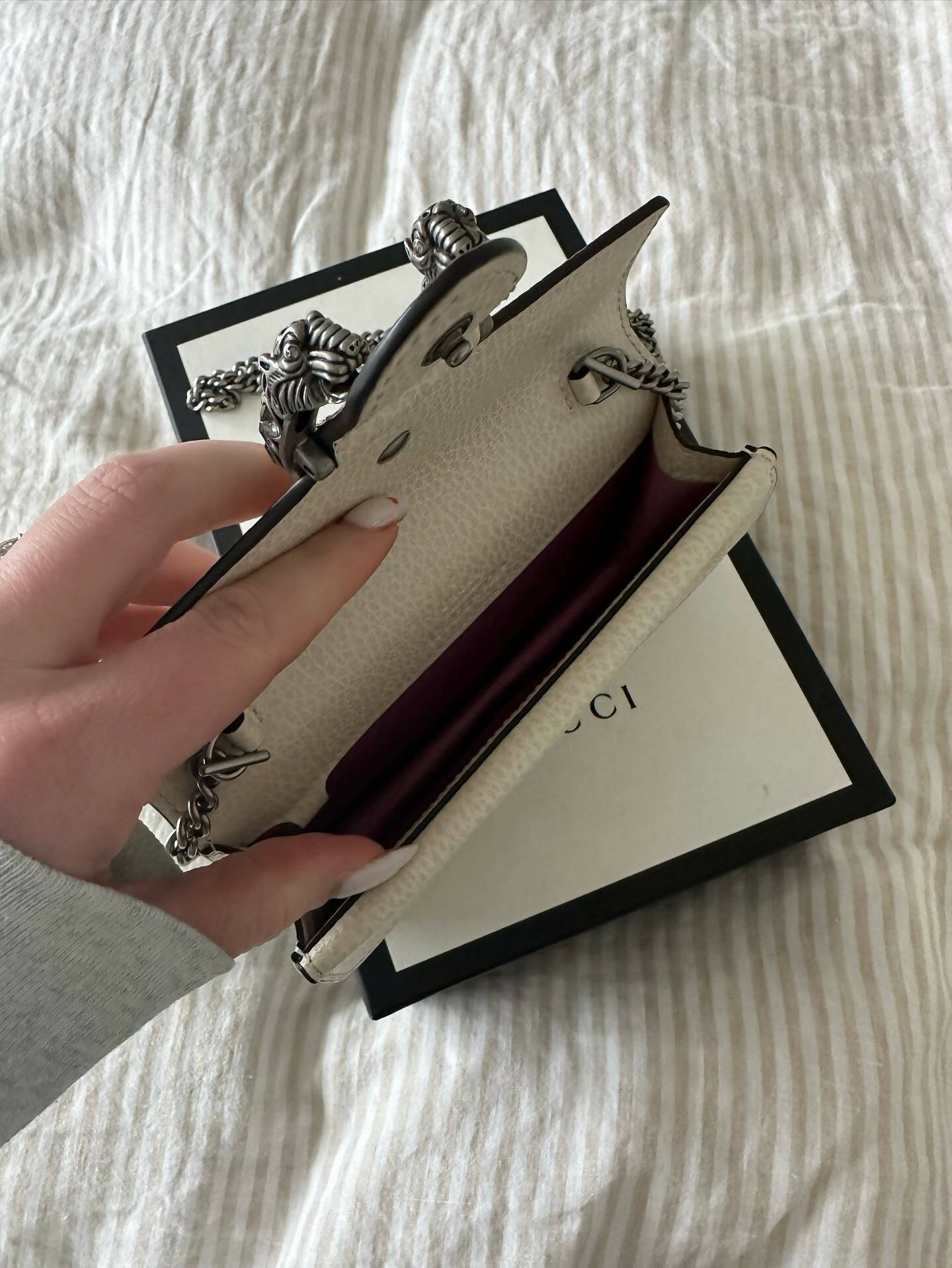 A hand opens an empty Gucci Dionysus Super Mini Crossbody Bag - FULL SET by EXCHANGE, featuring white textured leather and a chain strap, resting on a black and white Gucci box atop striped fabric.