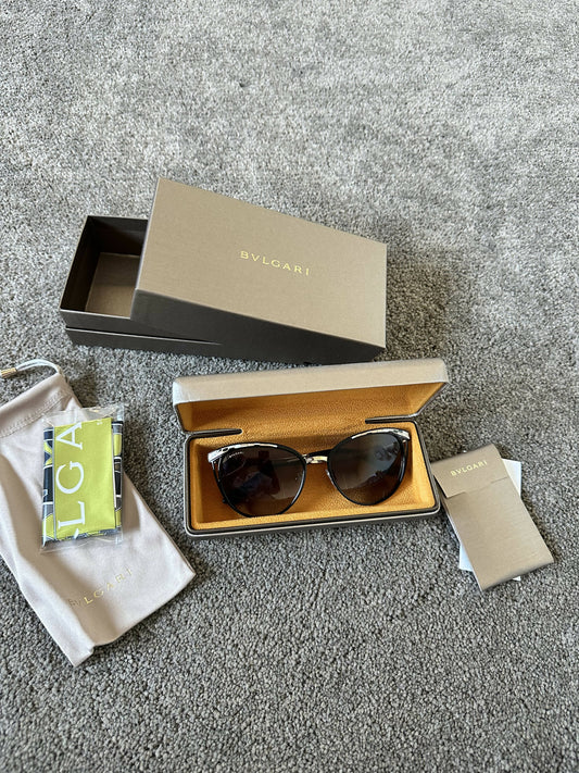 A pair of BVLGARI Serpenti Sunglasses by EXCHANGE with metal frames and gradient lenses are shown in an open case, alongside the branded box, cleaning cloth, booklet, and card, all placed on a gray carpet.