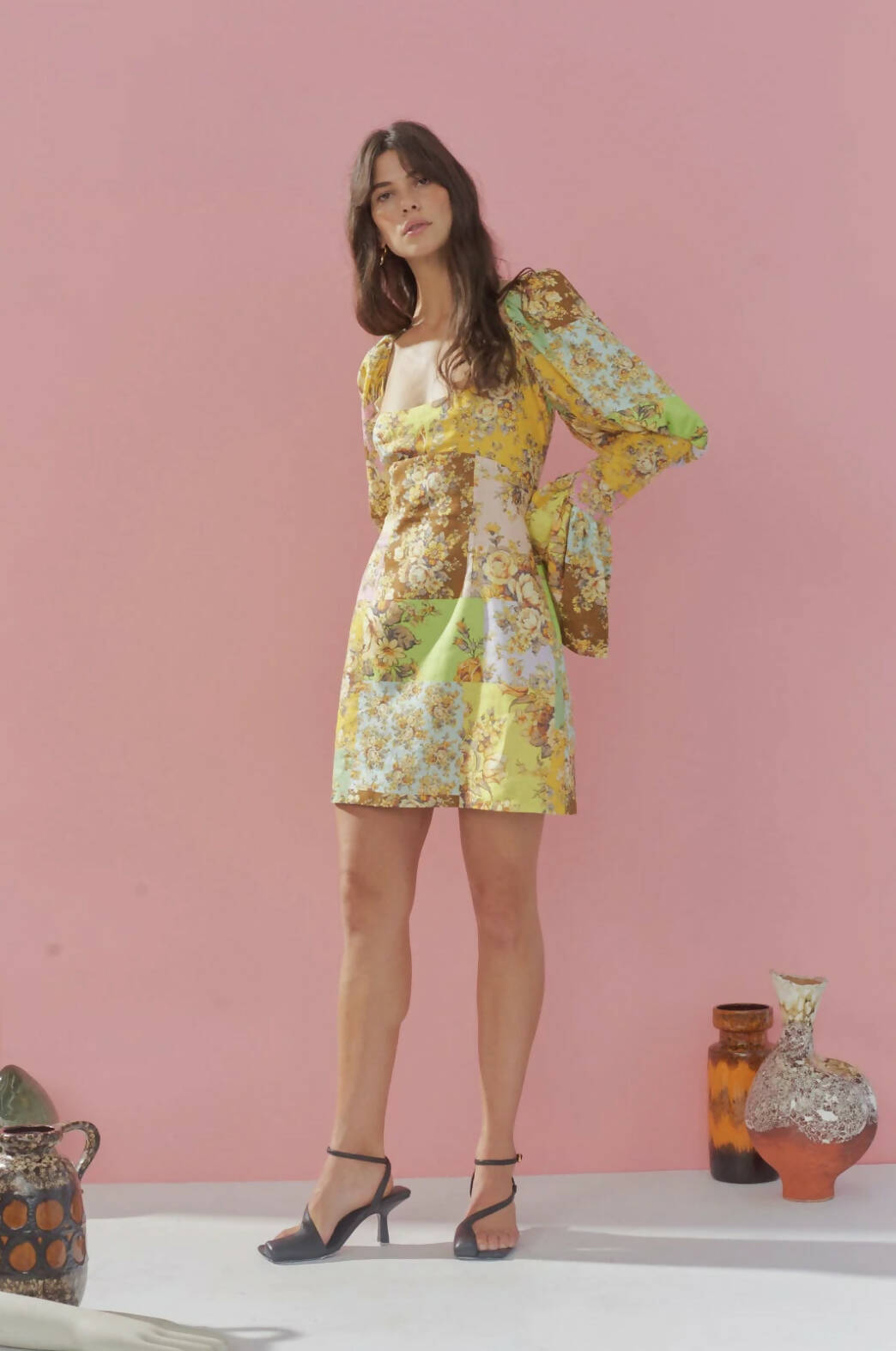 A woman wears the EXCHANGE Alemais Matilde Puff Sleeve Mini, a yellow floral dress with puff sleeves and fitted waist, styled with black heeled sandals. She stands against a pink wall surrounded by decorative ceramic vases.