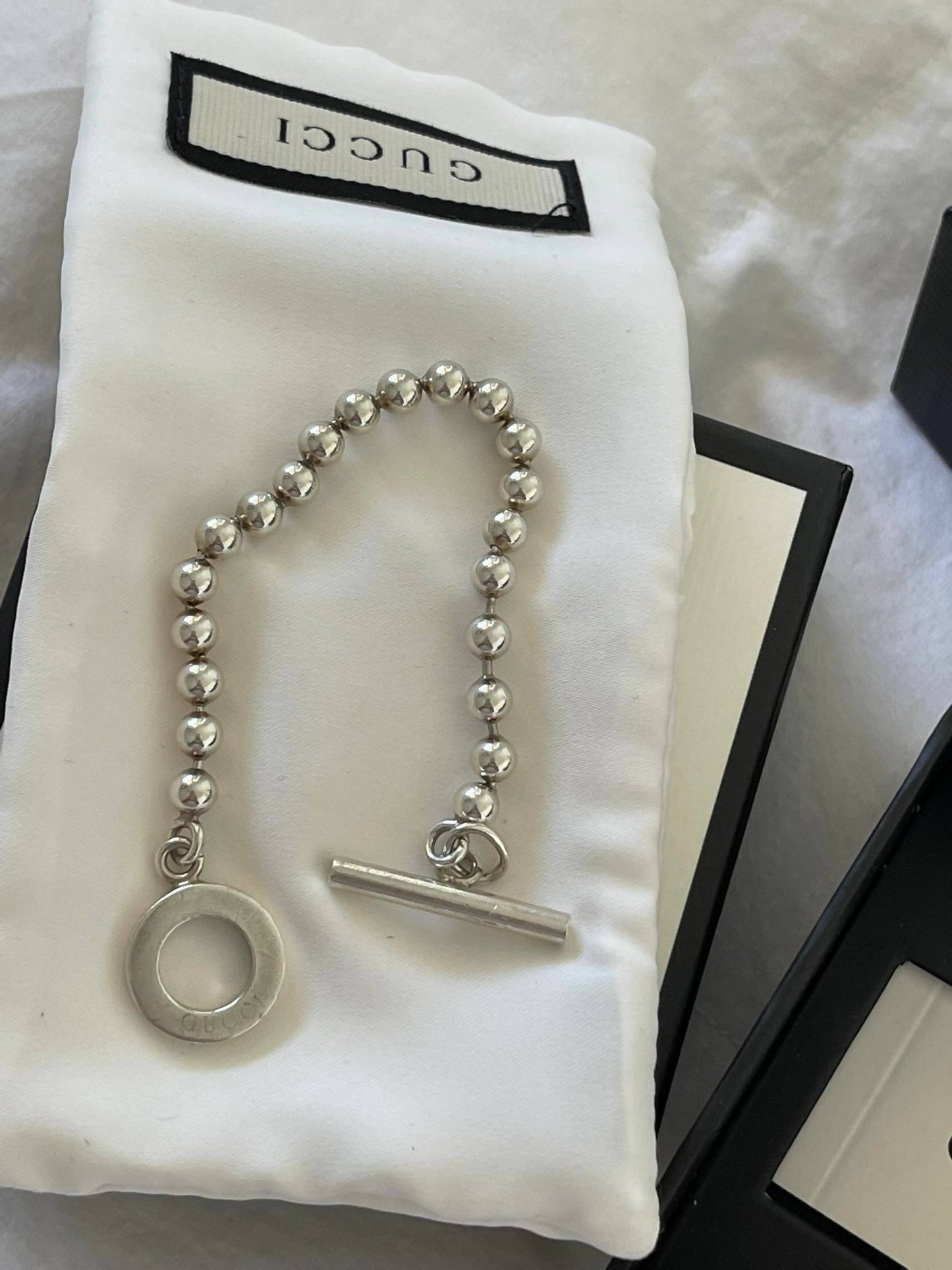 A small EXCHANGE Gucci Bracelet with beaded details, a circular charm, and bar clasp is displayed on a white cloth in a box, with the Gucci label visible at the top.