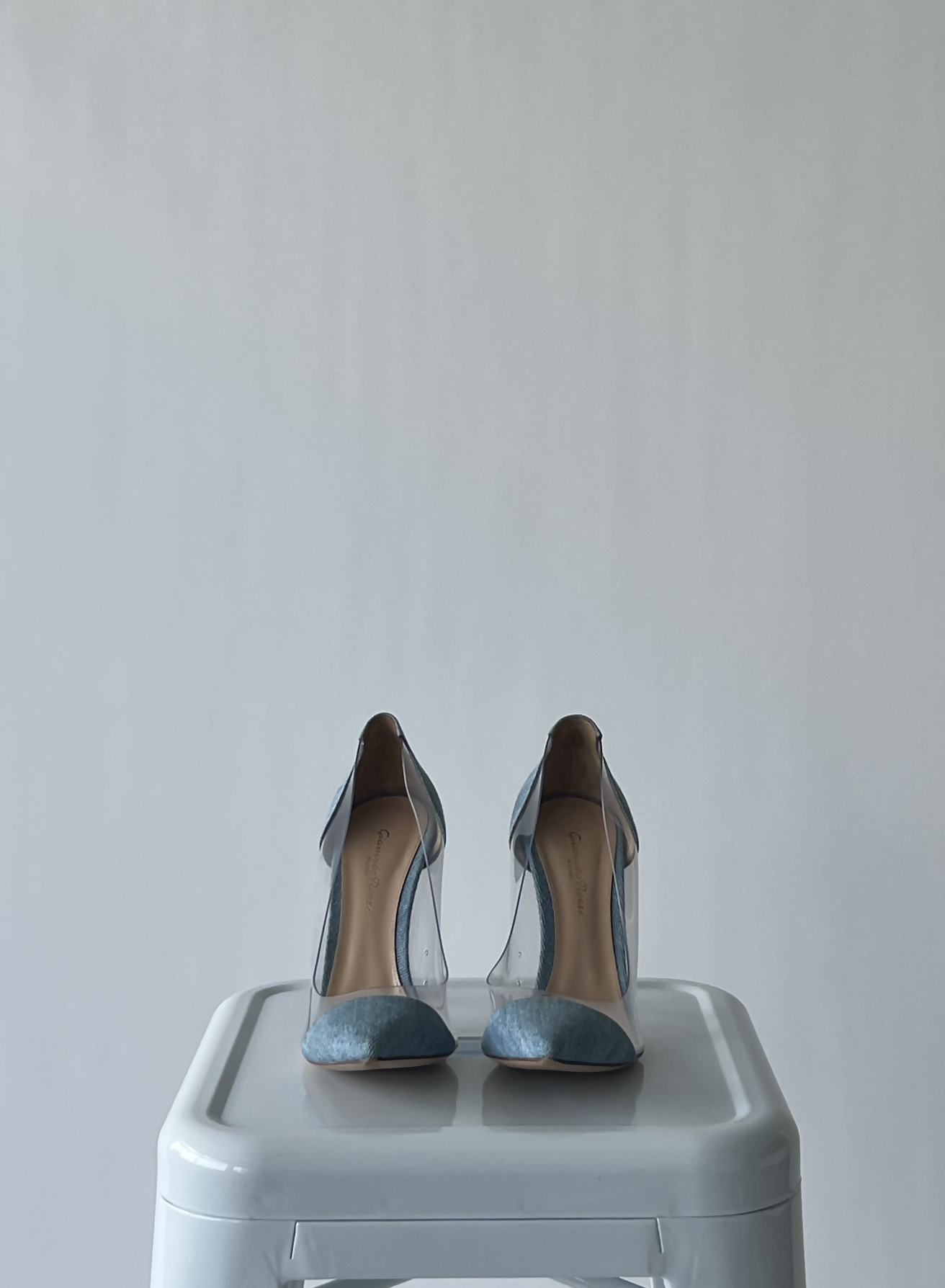 A pair of EXCHANGE Gianvito Rossi Light Blue Denim and PVC Plexi Pumps - FULL SET, in great condition, are placed side by side on a white stool against a plain light gray background.