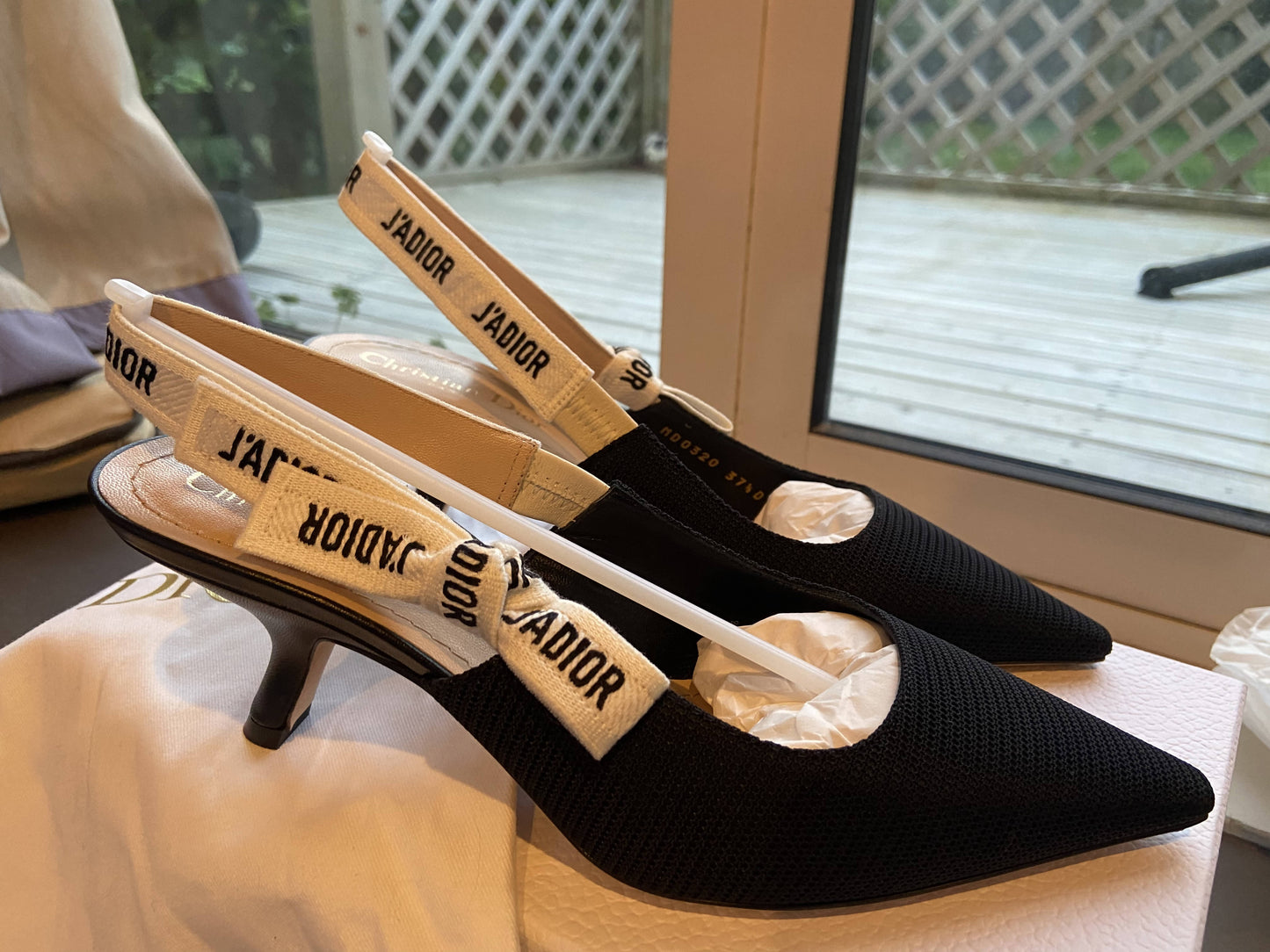A pair of Dior J’adior Slingback heels by EXCHANGE, size 37.5, black with white JADIOR straps, shown on a box beside a window overlooking an outdoor deck.