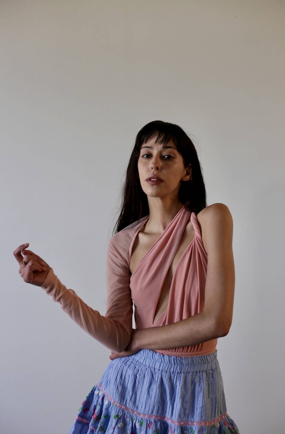 A woman with long dark hair poses against a plain background in an unworn pink Bao Tranchi Body Suit by EXCHANGE, featuring one long sleeve, paired with a blue patterned skirt. She looks at the camera with a neutral expression. Size 36.