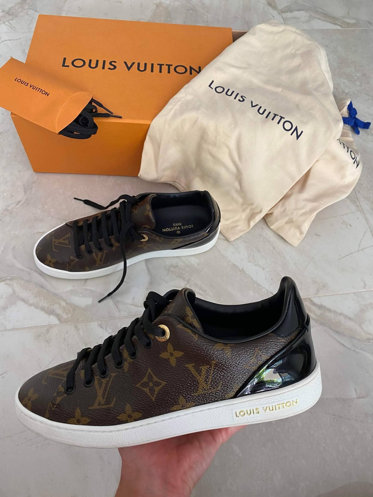 A hand holds an LV FRONTROW Sneaker, size 37, featuring the iconic monogram and LV laces. Another sneaker lies on marble, with an orange Louis Vuitton box and EXCHANGE-branded dust bag from exchange-store.com in the background.