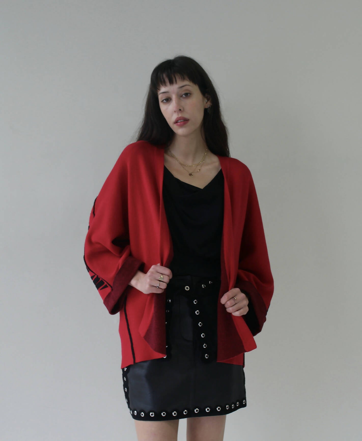 A woman with straight dark hair stands against a light background, wearing an EXCHANGE ARMANI EXCHANGE CARDIGAN in red over a black top and a black leather skirt with grommet details, looking at the camera with a neutral expression.
