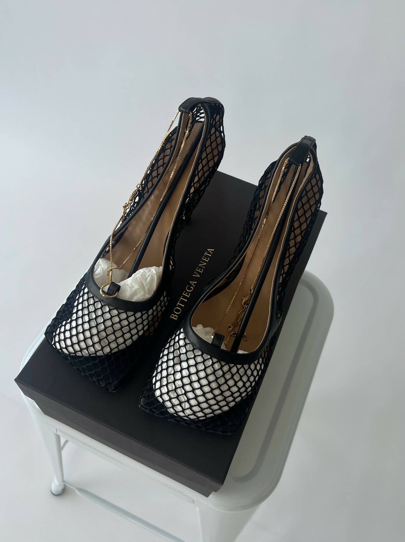 A pair of size 39 Bottega Veneta Mesh heels black by EXCHANGE, with pointed toes, displayed on a black box and placed on a white stool against a plain background.