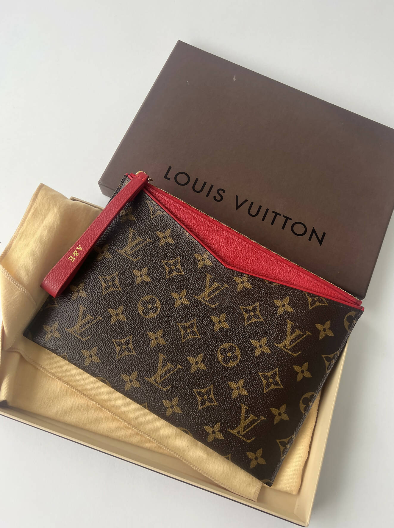 The Louis Vuitton Bicolor Monogram Pallas Pochette by EXCHANGE features a red zipper, wrist strap, and classic monogram. It rests on a beige cloth with receipts in an open box dustbag set, topped with a brown Louis Vuitton lid.