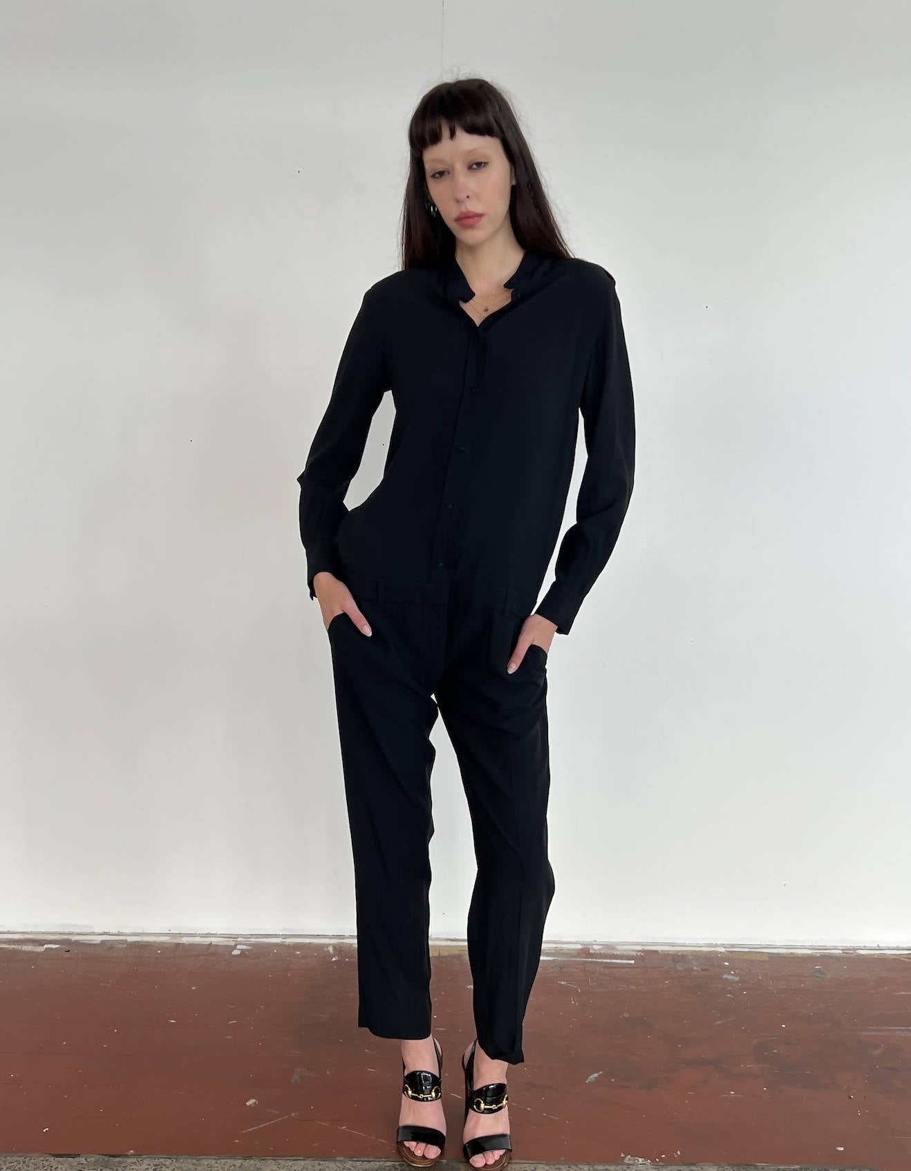 A person with long dark hair and bangs stands against a white wall, wearing a size 38 EXCHANGE Celine Jumpsuit and black open-toed heels. Their hands are in their pockets and they have a neutral expression.
