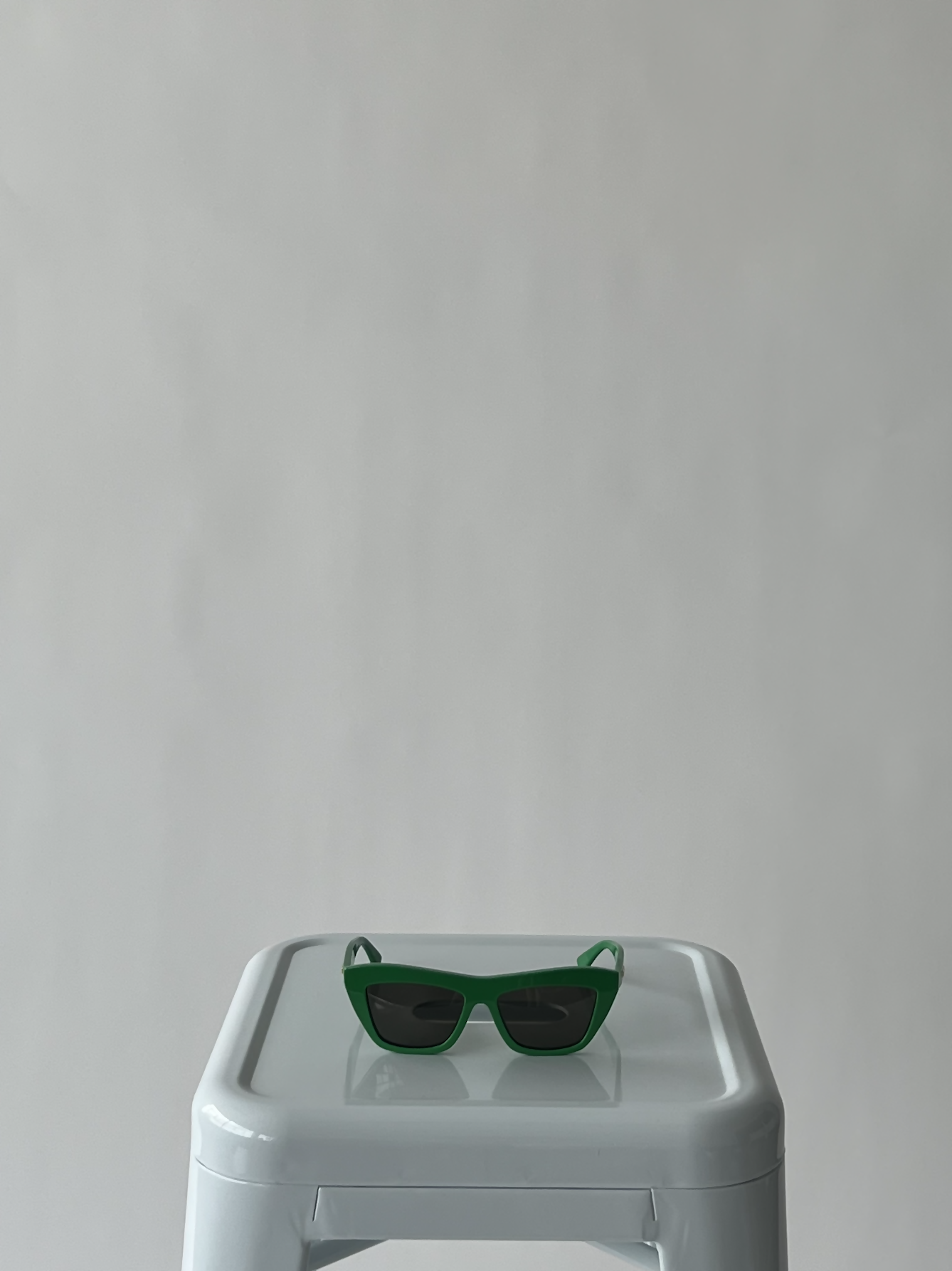 EXCHANGE Bottega Veneta New Entry Cat Eye Sunglasses rest atop a white plastic stool against a plain light gray background. Centered composition and soft lighting highlight the minimalistic elegance of these luxury sunglasses.