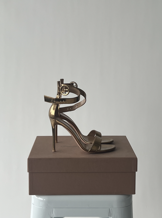 The Gianvito Rossi Gold Leather Portofino Ankle Strap Sandals by EXCHANGE rest on a closed brown box placed on a light gray stool against a plain light background.