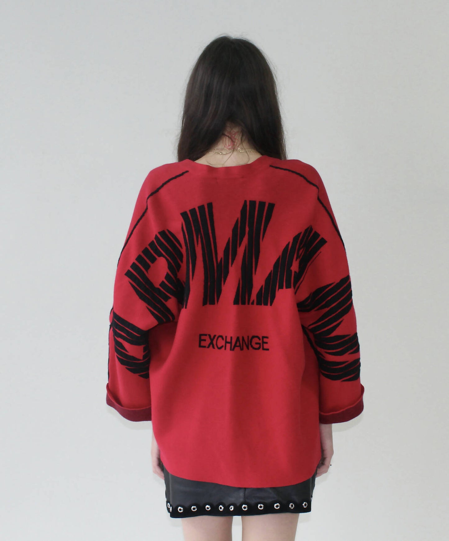 A person with long dark hair, seen from behind, wears a loose red ARMANI EXCHANGE CARDIGAN with bold black graphic lines and "EXCHANGE" on the back, paired with a short black skirt featuring grommet details.