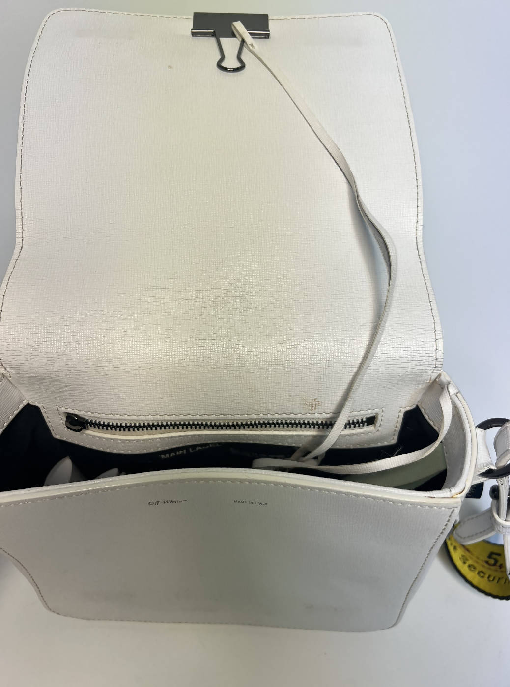 The EXCHANGE Off-White diagonal stripe crossbody bag features a white textured design, open flap with black binder clip, internal zip pocket, yellow strap, and black text detail.