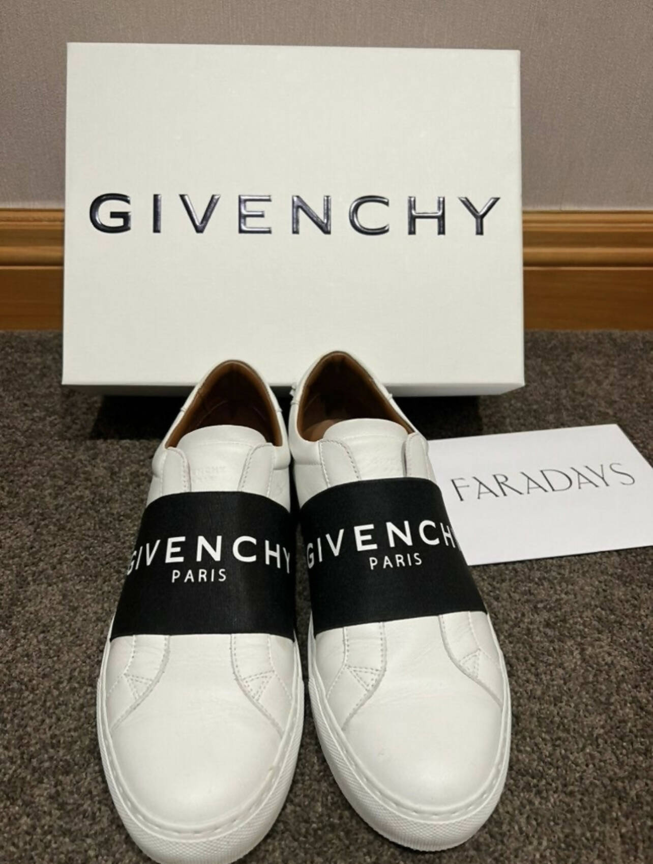 A pair of white EXCHANGE Givenchy Urban Street Sneakers in size 40 EU, featuring black brand-name bands, is displayed with a Givenchy shoebox and Faradays card on a carpeted floor.