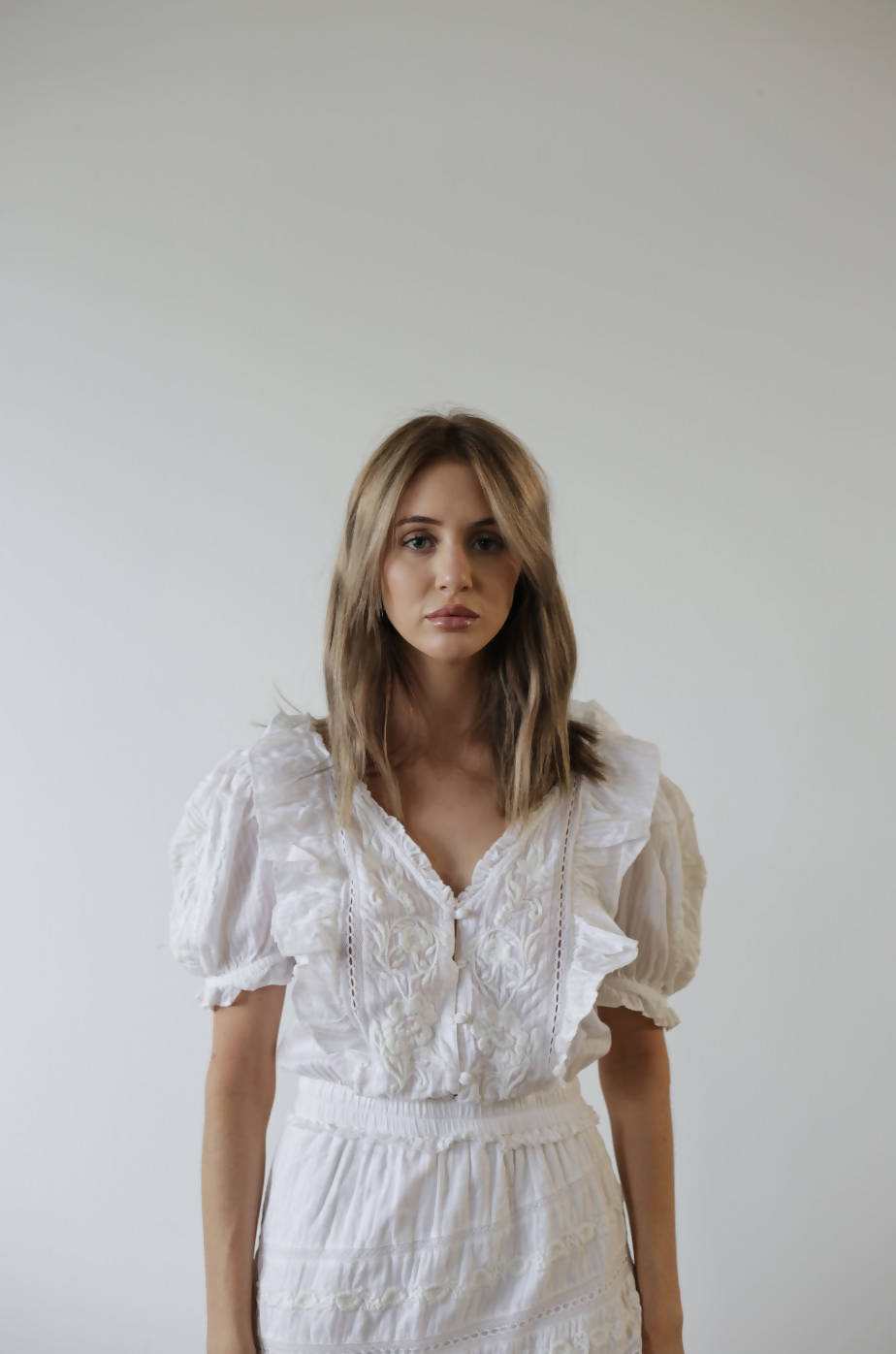 A woman with light brown hair wears an EXCHANGE LoveShackFancy White Dress with ruffled short sleeves and skirt, standing against a plain light-colored background and looking at the camera.