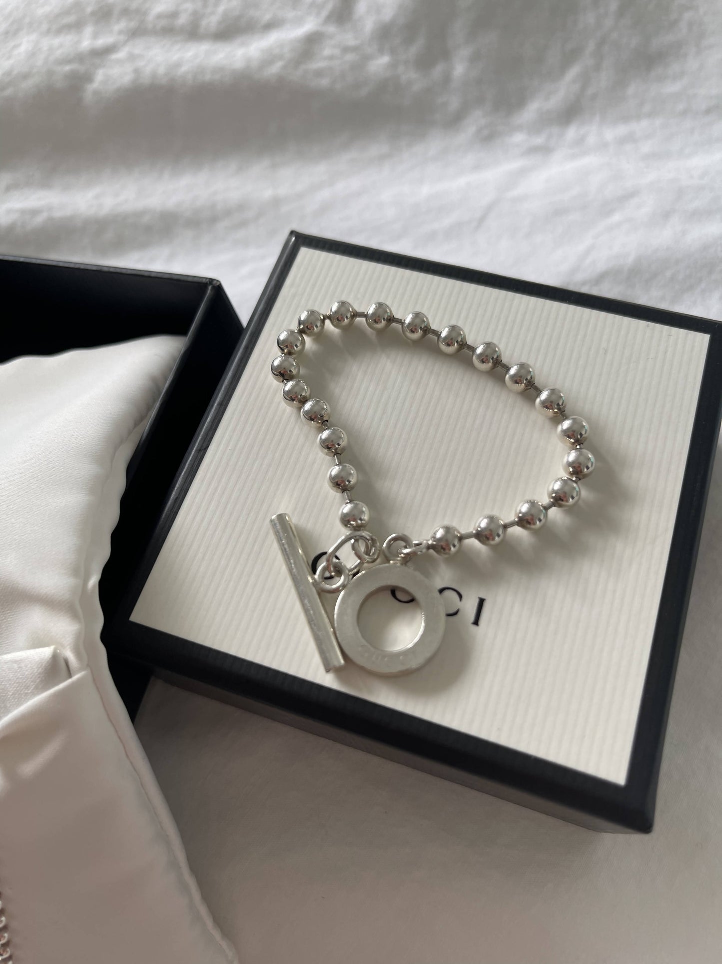The EXCHANGE Gucci Bracelet, featuring sterling silver round and bar charms, is showcased on a white box with black edges beside a satin pillow inside an open gift box—an elegant piece for a refined touch to any outfit.