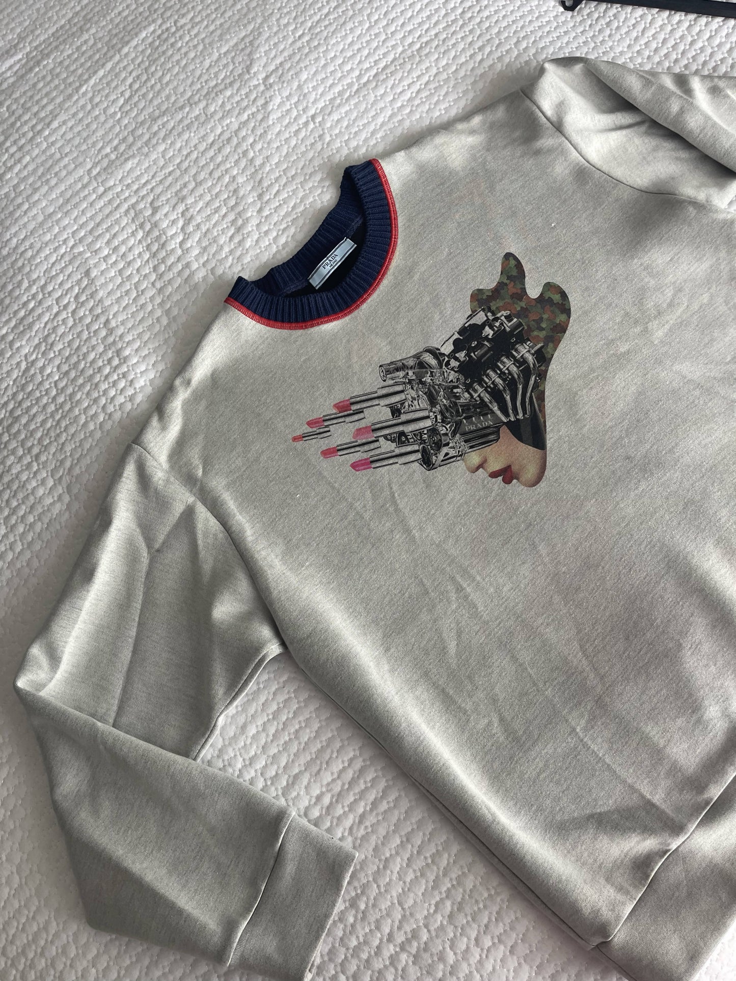 The PRADA 2X4 Collaboration Jumper, Grey by EXCHANGE features a graphic collage of a face and industrial structure on its upper front. It boasts a red and black collar, with the artwork set against a textured white surface.