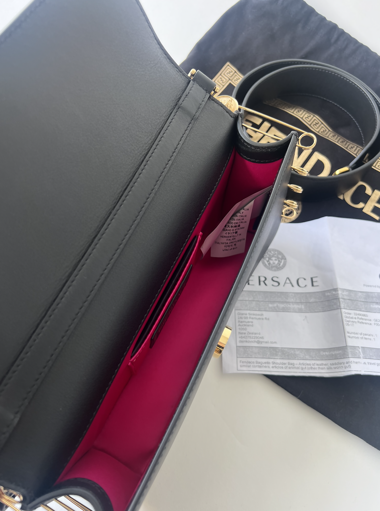 An open black FENDI X Versace Fendace Baguette Shoulder Bag by EXCHANGE, with a bright pink interior, sits beside a black dust bag and authentication document on a white surface, highlighting iconic Italian style.