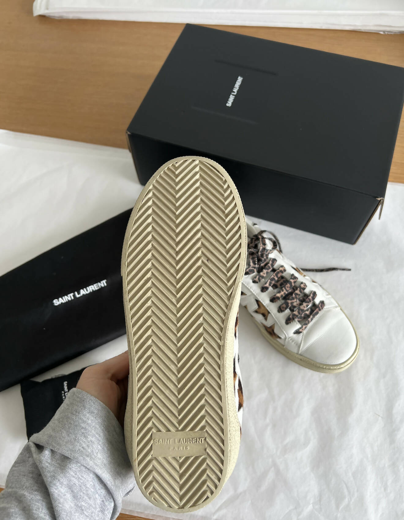 A hand holds up a Saint Laurent Court Classic Sneaker in Leopard/Cheetah, highlighting the textured sole. Another sneaker, shoebox, and Saint Laurent dust bag are displayed—available now at EXCHANGE in the Auckland Region.