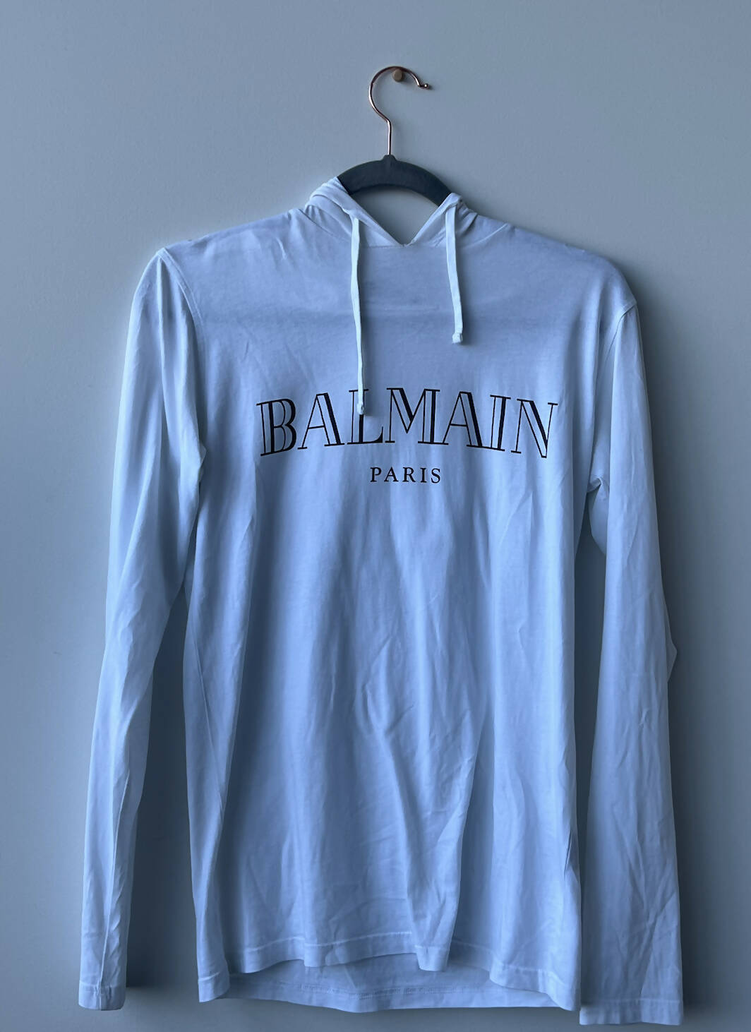 A white Balmain Long Sleeve Hoodie 2017 by EXCHANGE hangs on a black hanger against a gray wall, made from 100% cotton and featuring "BALMAIN" in bold black letters with "PARIS" below.
