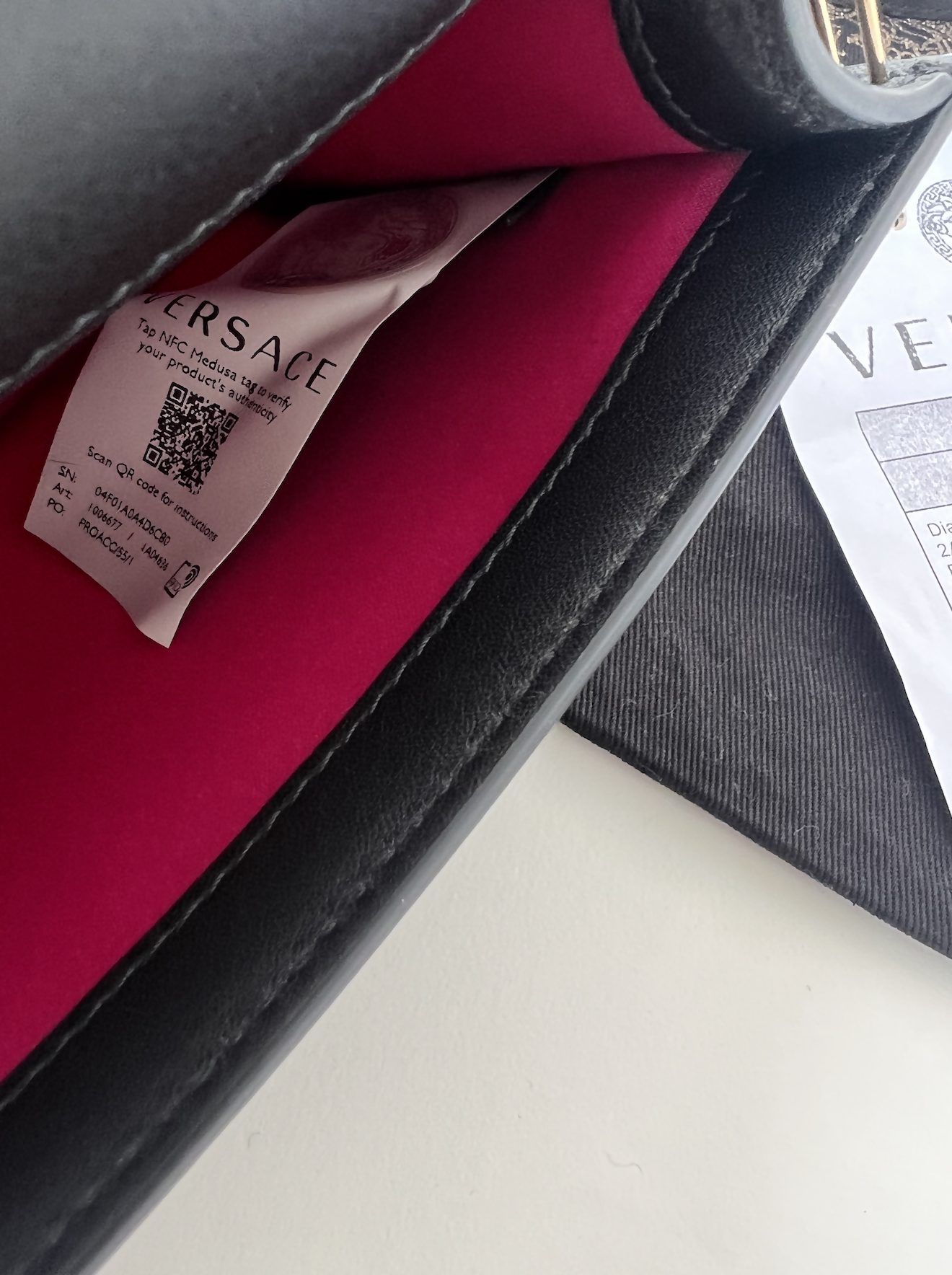 Close-up of the EXCHANGE FENDI X Versace Fendace Baguette Shoulder Bag’s interior, featuring a pink lining, branded tag with QR code, and part of an authenticity card on a dark fabric surface.