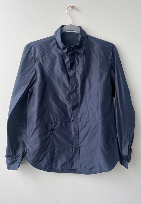 The EXCHANGE Christian Dior Shirt in navy silk, featuring long sleeves, a collar, straight hem, and subtle front stitching, hangs on a copper hanger against a white wall—an understated luxury piece.