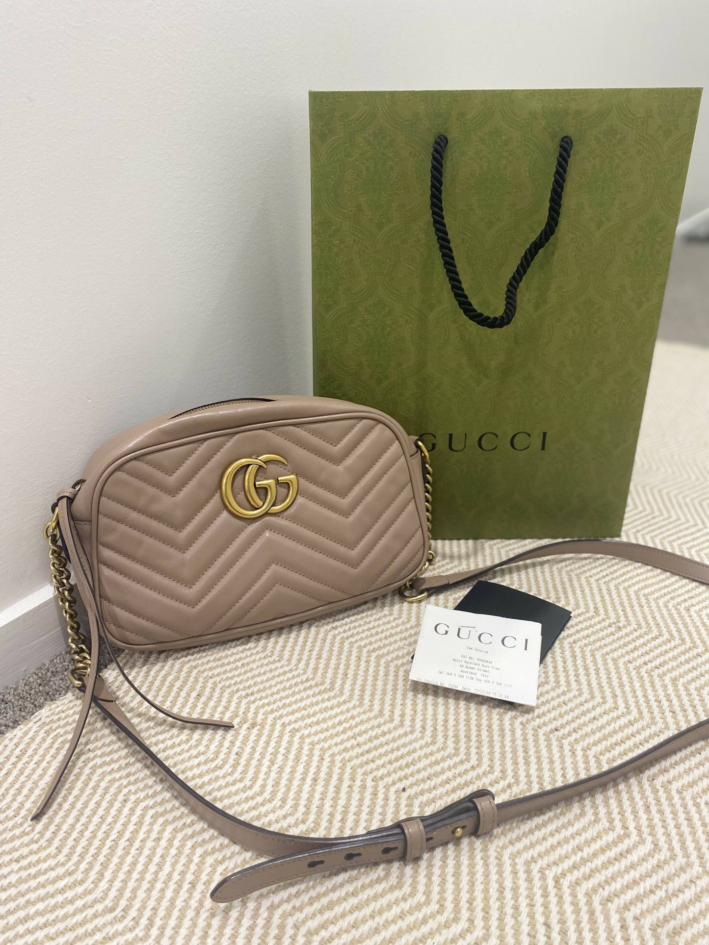 A Gucci Marmont Matelasse Small Dusty Pink bag by EXCHANGE is displayed before a green Gucci paper bag, with a receipt and small black pouch on a beige-patterned surface.