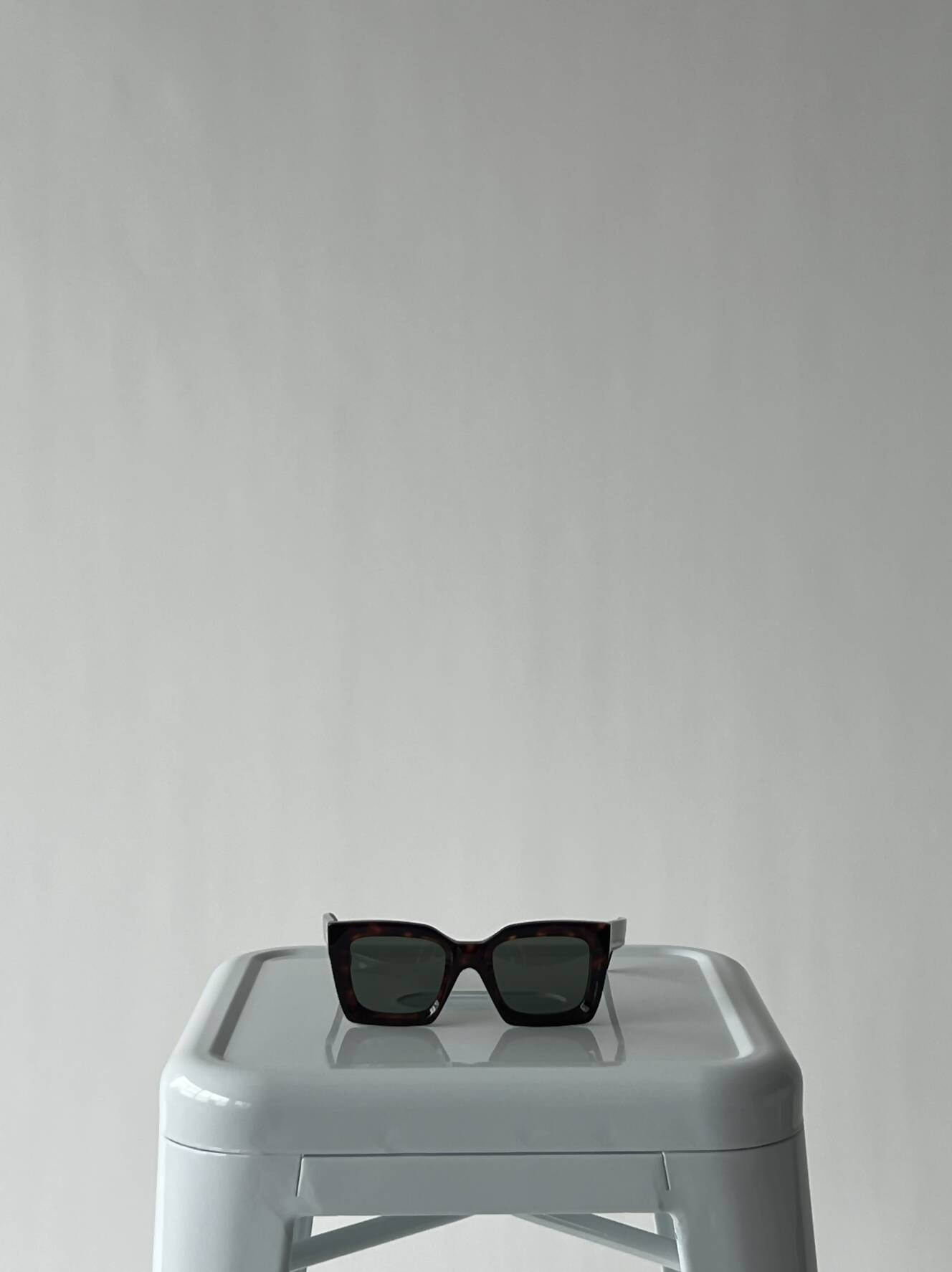 A pair of EXCHANGE Celine Glasses - CL40130I with a black rectangular acetate frame sits atop a light gray plastic stool against a plain, light background.