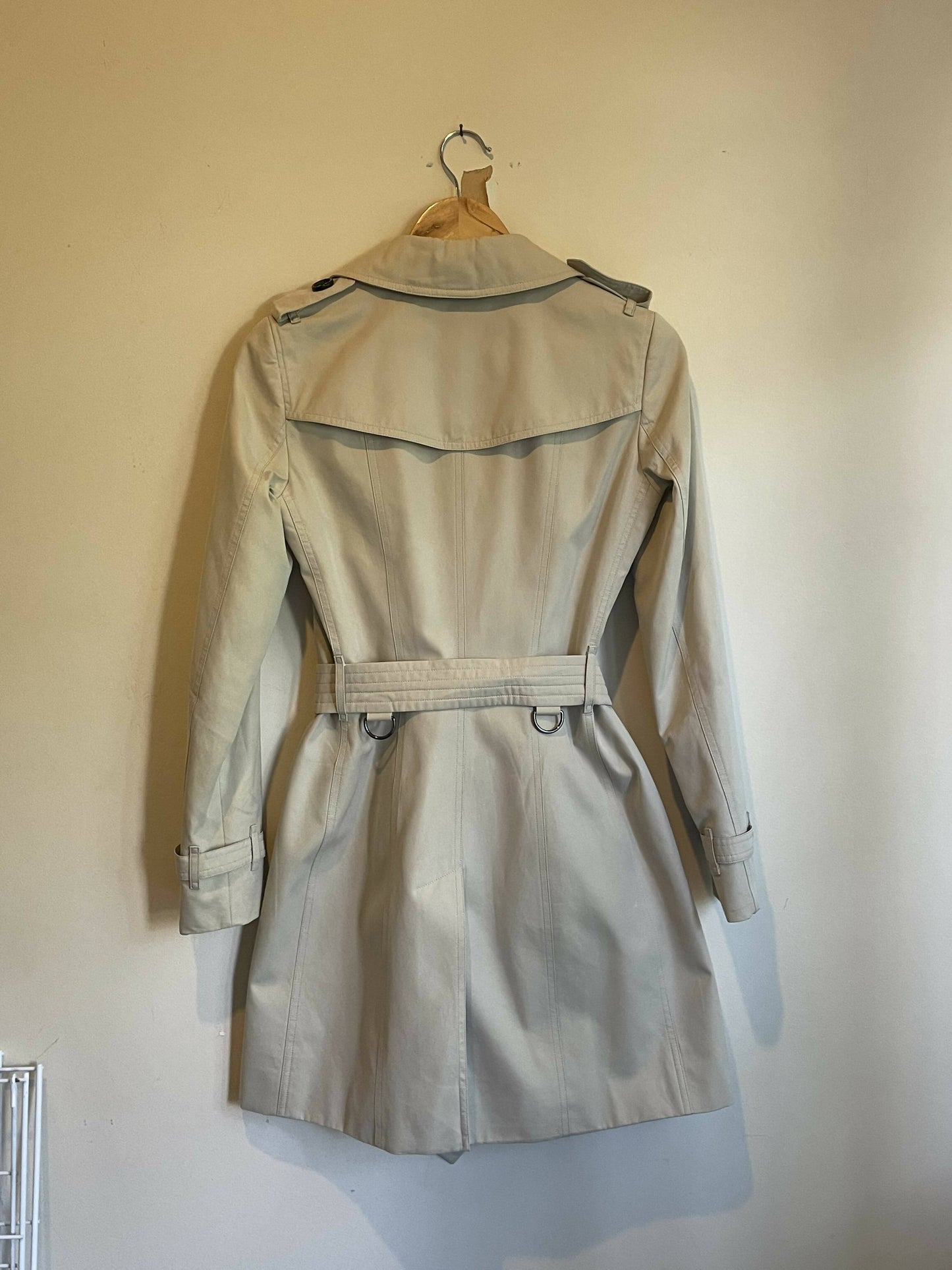 The EXCHANGE Burberry - Mid-length Chelsea Heritage Trench Coat UK Size 6, in beige gabardine with a belt, shoulder epaulettes, long sleeves, and vintage check lining, hangs on a wooden hanger against a light wall.