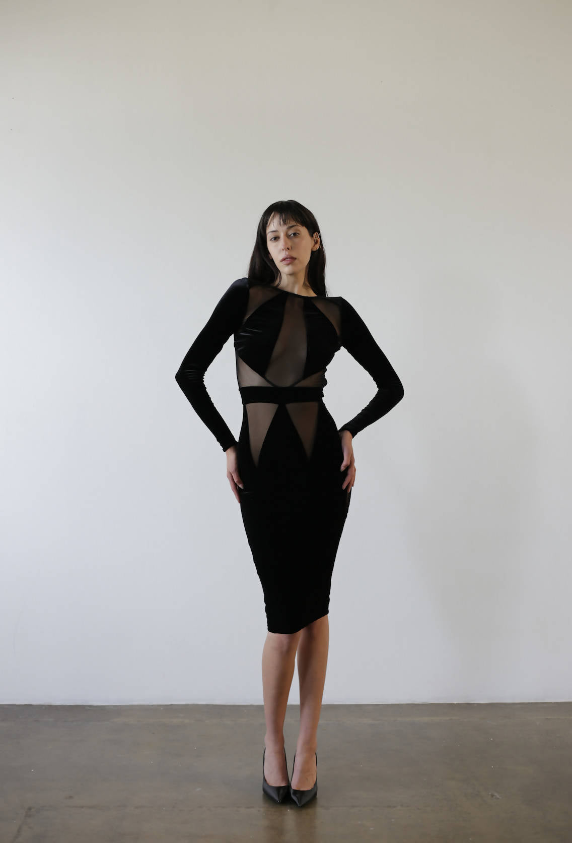 In a minimalist room, a person with long dark hair poses elegantly against a light wall in an EXCHANGE Bao Trenchi Dress, featuring black and sheer mesh with long sleeves, paired with black heels.