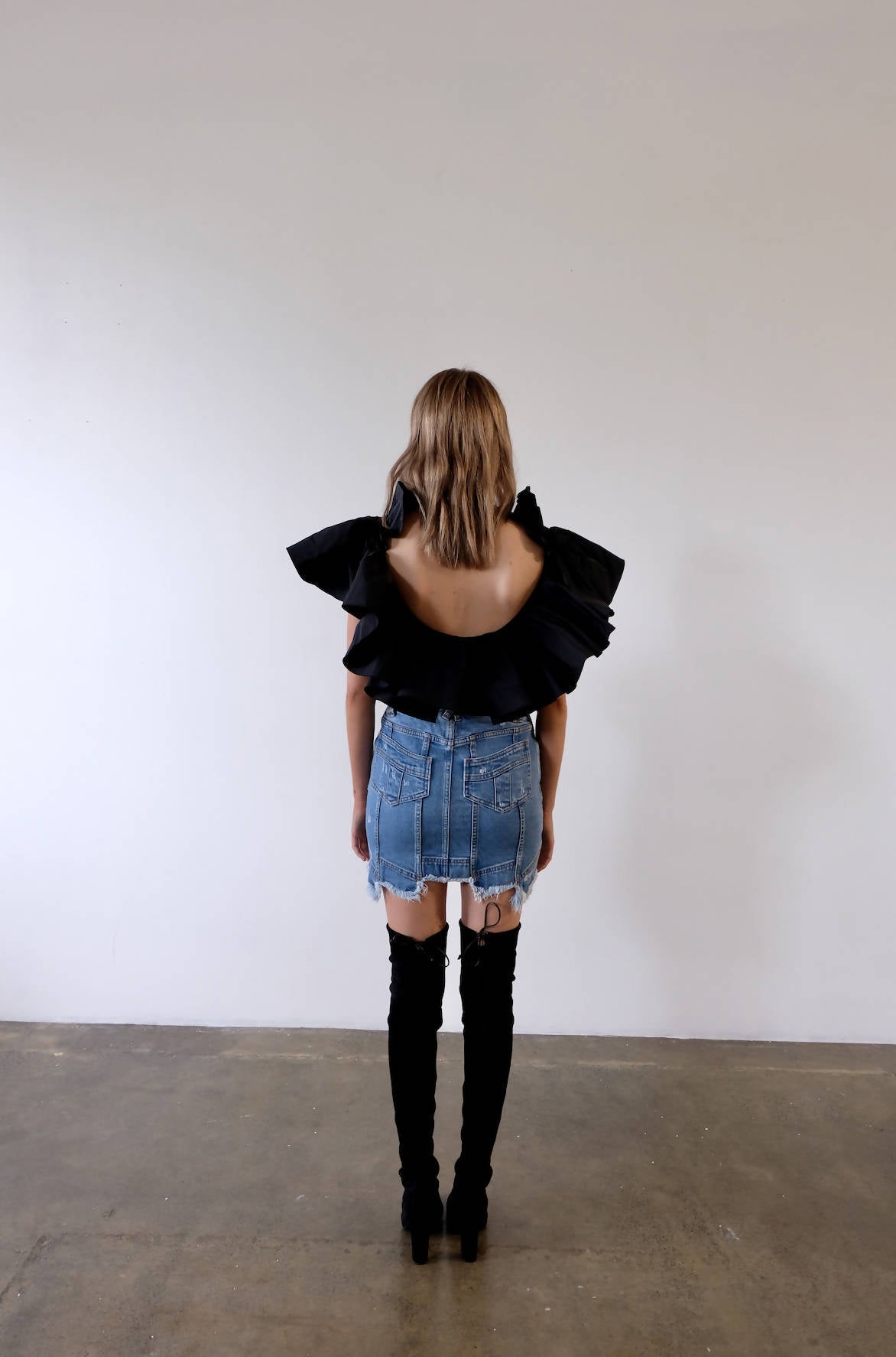 A woman faces a white wall, wearing an EXCHANGE Alexander Macqueen Body Suit, a frayed denim mini skirt, and black over-the-knee boots in size 36 from our exchange-store. She stands on a concrete floor.