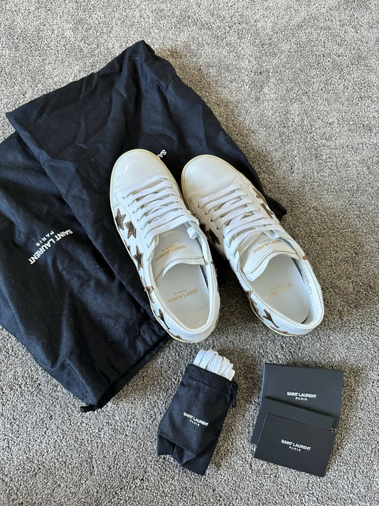 A pair of EXCHANGE Saint Laurent Court Classic Sneakers in white leather with silver star patches is displayed on a carpet, accompanied by black dust bags, extra white laces in a pouch, and several Saint Laurent cards.