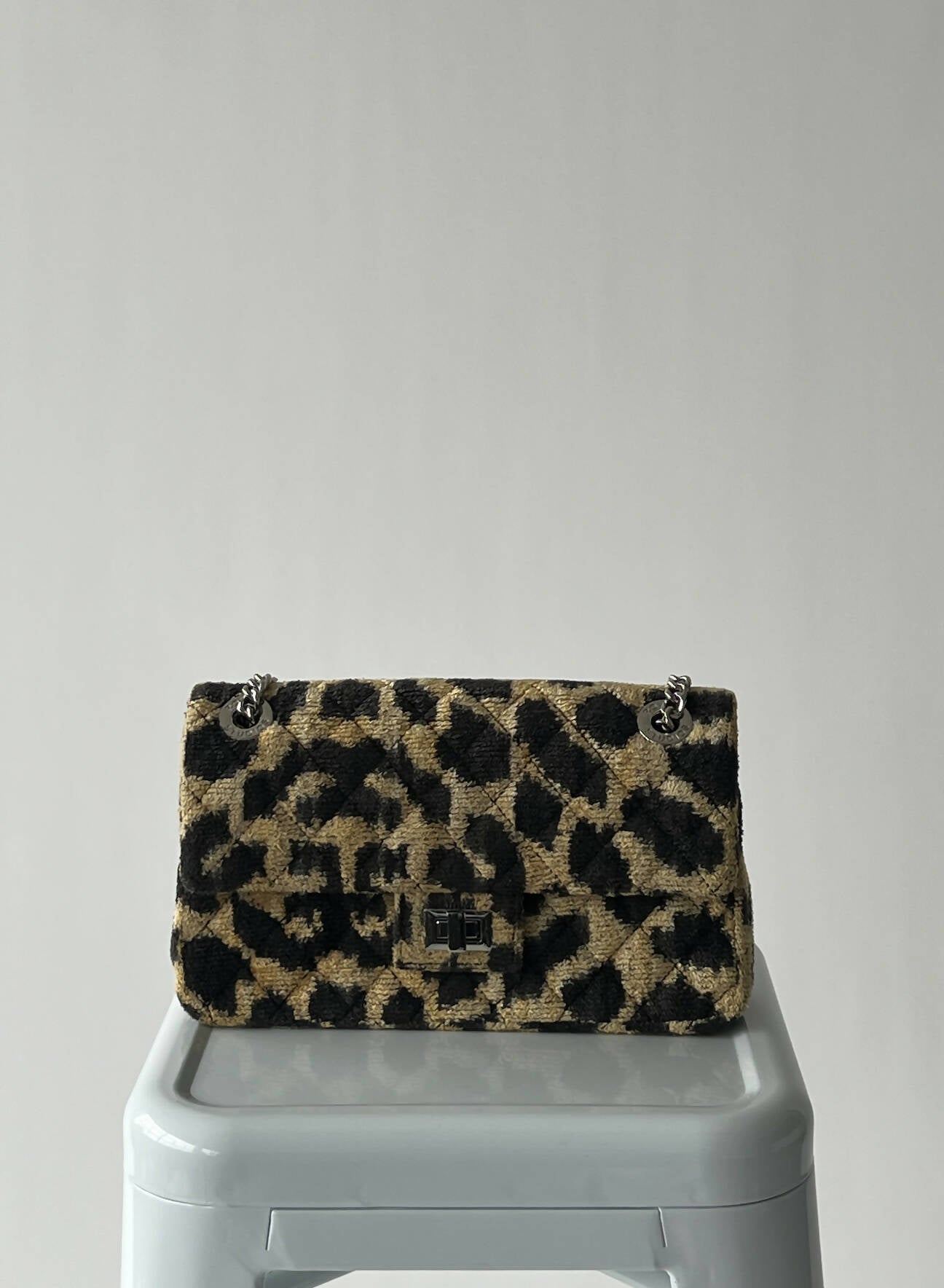 A used Rough Studios Bag by EXCHANGE in black and brown leopard print with silver chain straps and metal clasp rests on a light gray stool. This stylish handbag includes a dustbag for added convenience.