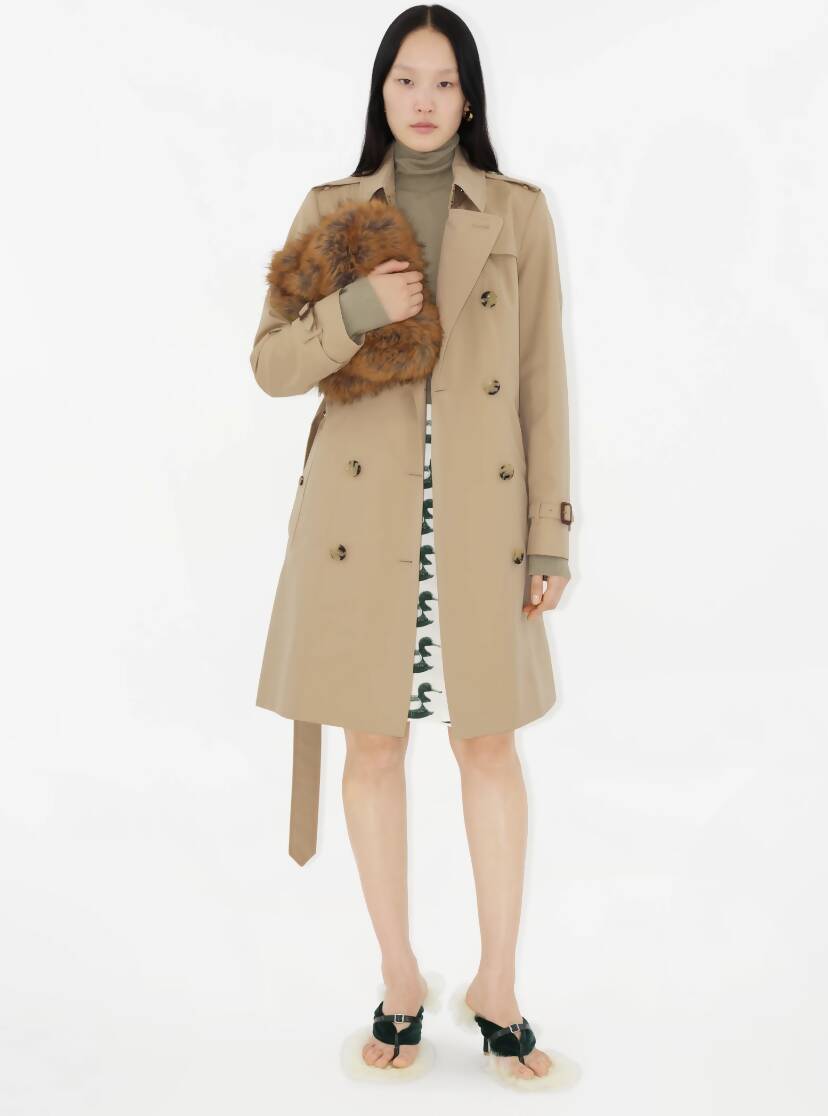 A woman with long black hair stands against a white background, wearing an EXCHANGE Burberry Mid-length Chelsea Heritage Trench Coat in UK Size 6 over a green turtleneck and patterned white skirt, accessorized with a fluffy brown purse and fur-accented open-toed shoes.
