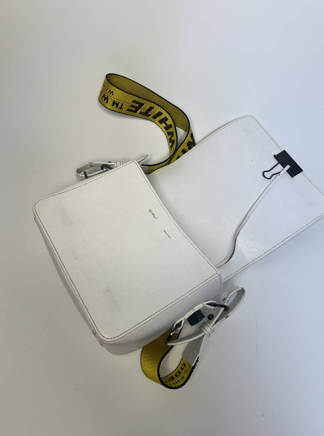 A white EXCHANGE Off-White diagonal stripe crossbody bag with a yellow industrial strap and black binder clip closure sits slightly open on a white surface, revealing its interior flap.