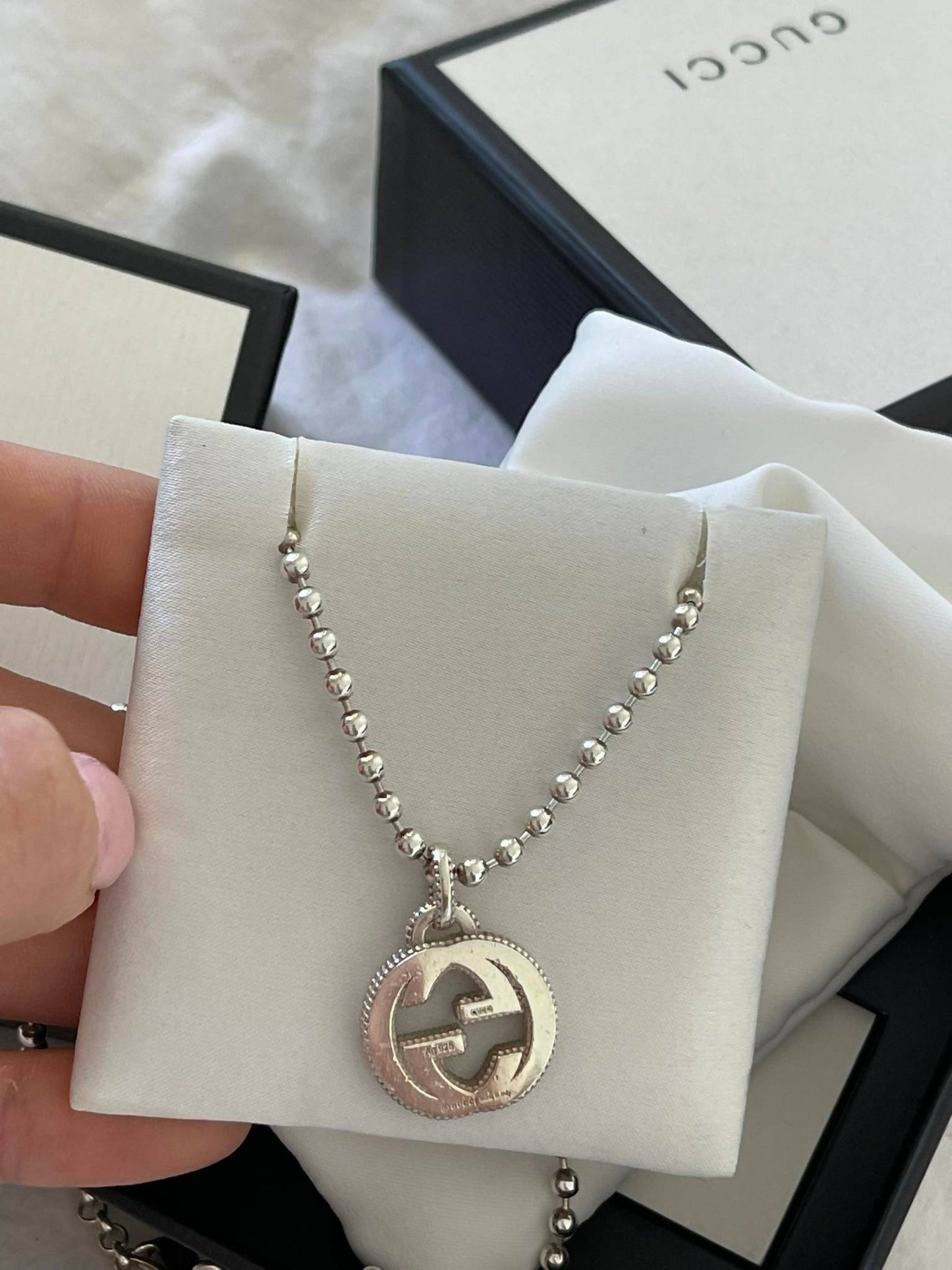 A hand holds the EXCHANGE Gucci Bracelet with a circular pendant featuring the interlocking logo, displayed on a white padded card and Gucci packaging in the background.