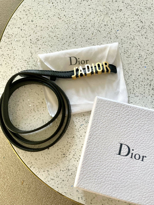 A Christian Dior J’adior Waist Belt by EXCHANGE with gold lettering lies on a white pouch next to a textured Dior box atop a speckled surface.