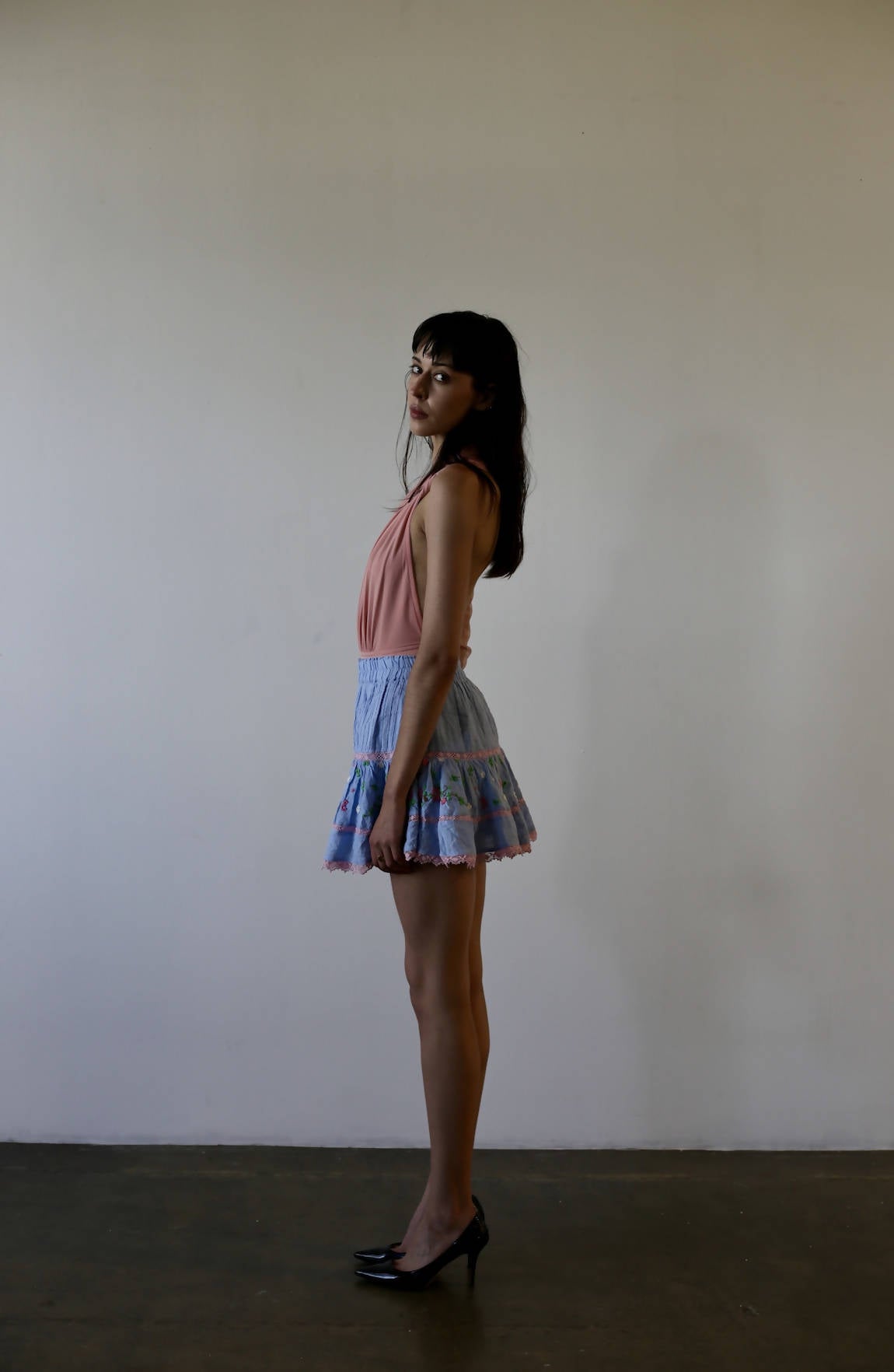 A woman stands in profile against a white wall, wearing an unworn sleeveless pink Bao Tranchi Body Suit by EXCHANGE, a blue floral ruffled skirt (size 36), and black heels. Soft lighting creates a gentle shadow on the wall.