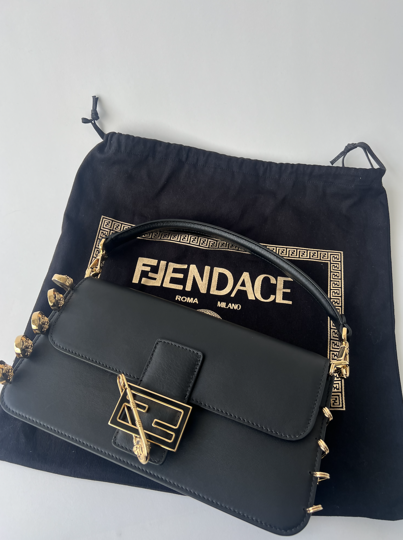 The FENDI X Versace Fendace Baguette Shoulder Bag by EXCHANGE features black leather, a gold clasp, and chain accents, resting on a Fendace ROMA MILANO dust bag—a stunning showcase of Italian fashion and luxury.