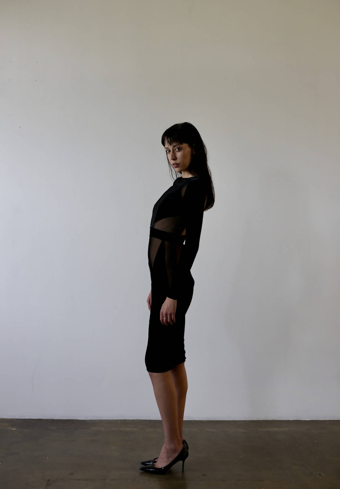 A woman models the Bao Trenchi Dress by EXCHANGE, standing sideways against a light gray backdrop in a black, semi-transparent mesh dress and black velvet heels, with long dark hair. The image exudes a minimalist and fashion-forward aesthetic.