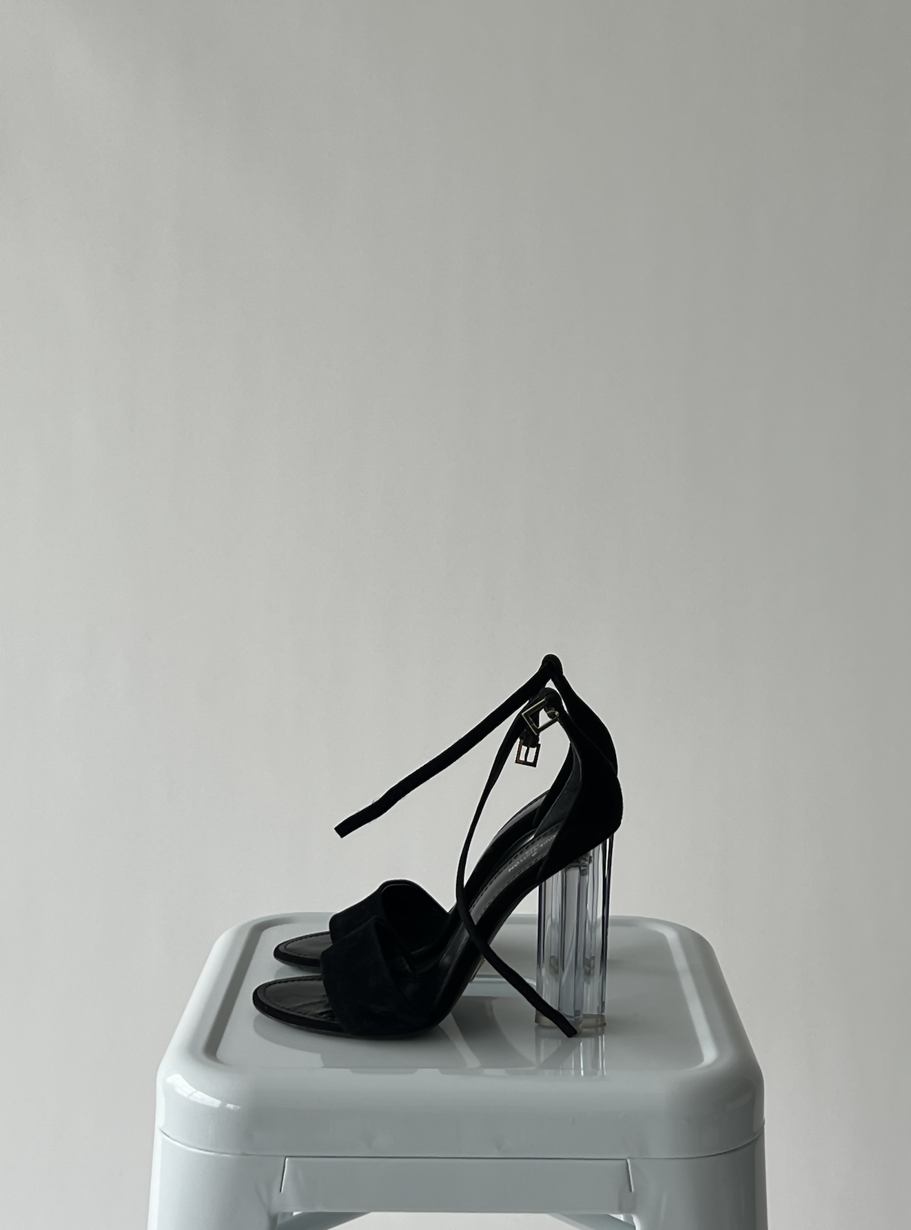 The Louis Vuitton Crystal Flower Sandals by EXCHANGE, featuring black uppers, a clear chunky heel, and an ankle strap, are displayed on a white stool against a plain light gray background.