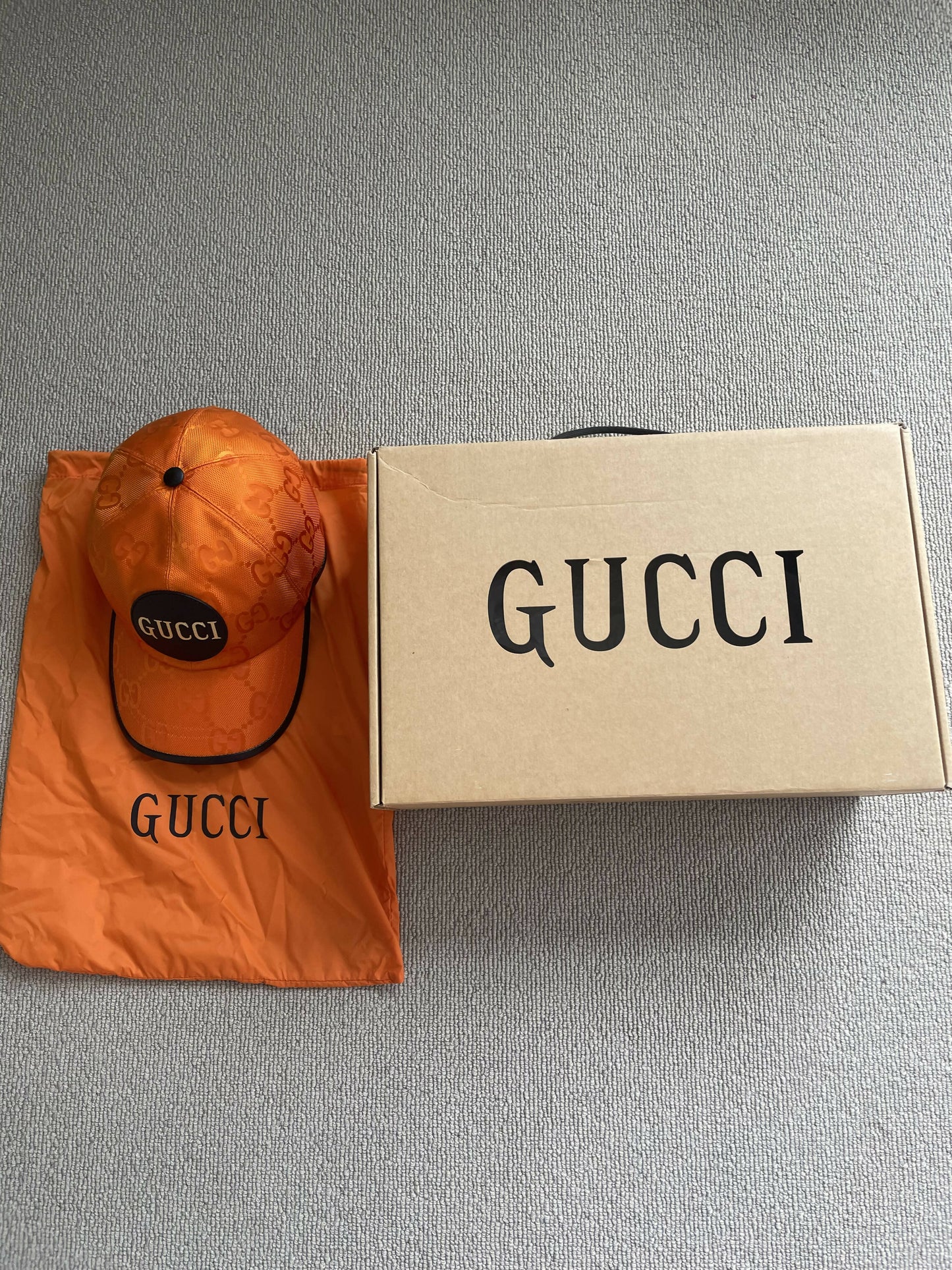 The EXCHANGE Gucci Off The Grid GG Supreme Canvas Baseball Cap and matching dust bag are displayed beside a beige Gucci box on a light gray textured surface, highlighting the signature collection.