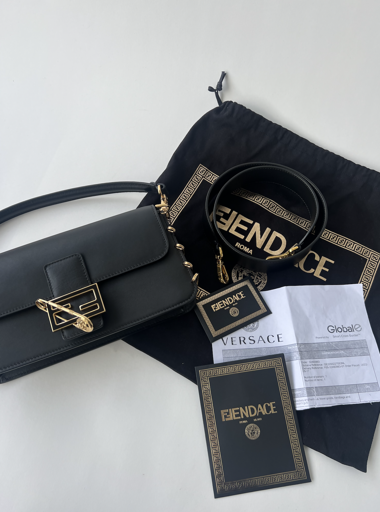 A black FENDI X Versace Fendace Baguette Shoulder Bag by EXCHANGE with a matching belt and gold accents is displayed on a black Fendace dust bag, accompanied by branded tags, a Versace card, and paperwork on a white surface.