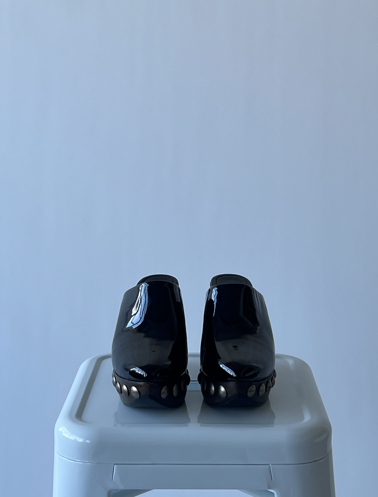 A pair of EXCHANGE Alaïa Le Sabot leather and wood pumps with metallic-studded soles sits side by side on a white stool against a plain light background.