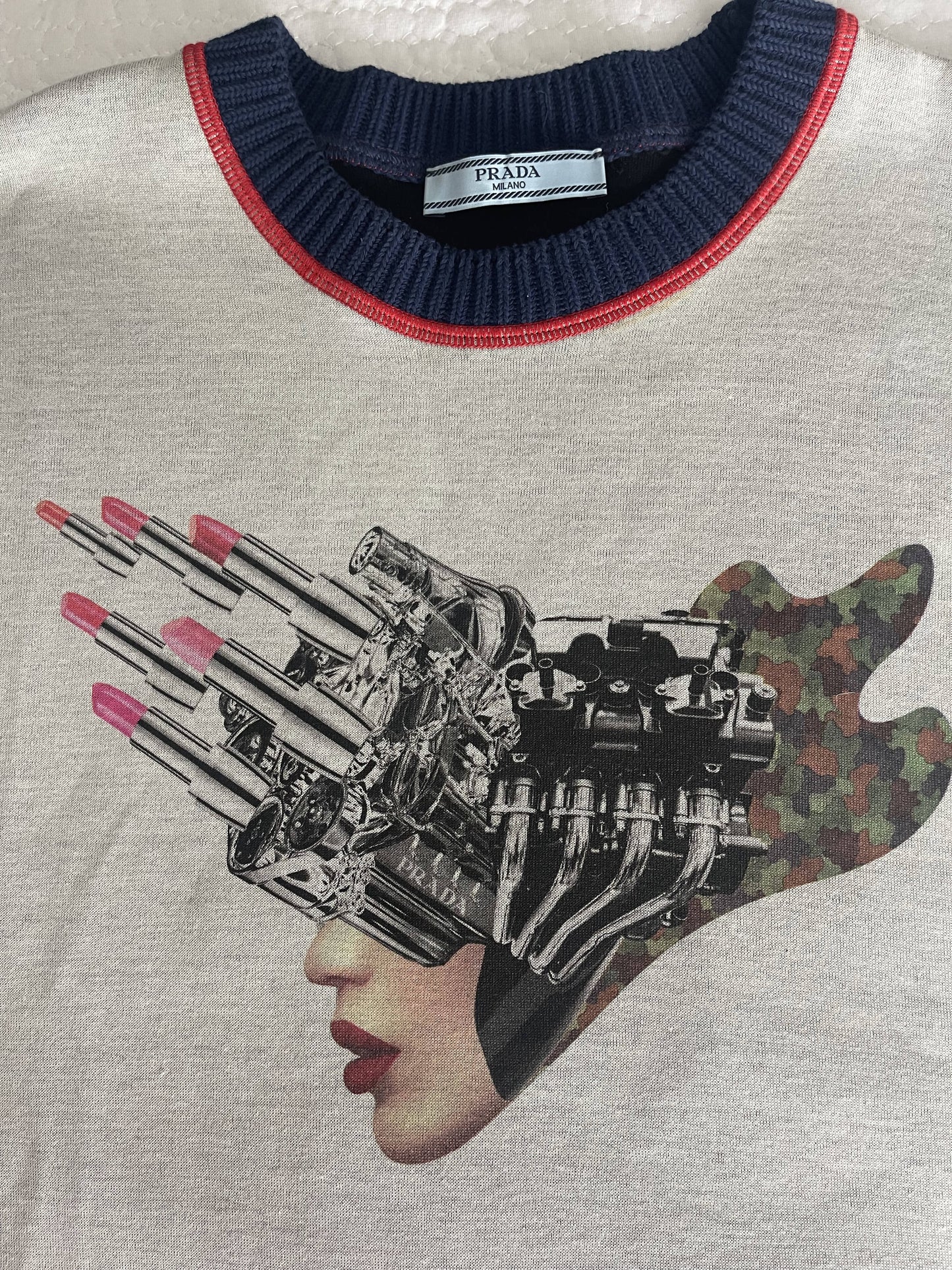 The PRADA 2X4 Collaboration Jumper in grey by EXCHANGE features a surreal lipstick face design with a womans visage and red lips merging into a metallic engine, lipsticks projecting camo patterned shadows, and finished with an eye-catching blue and red crewneck collar.