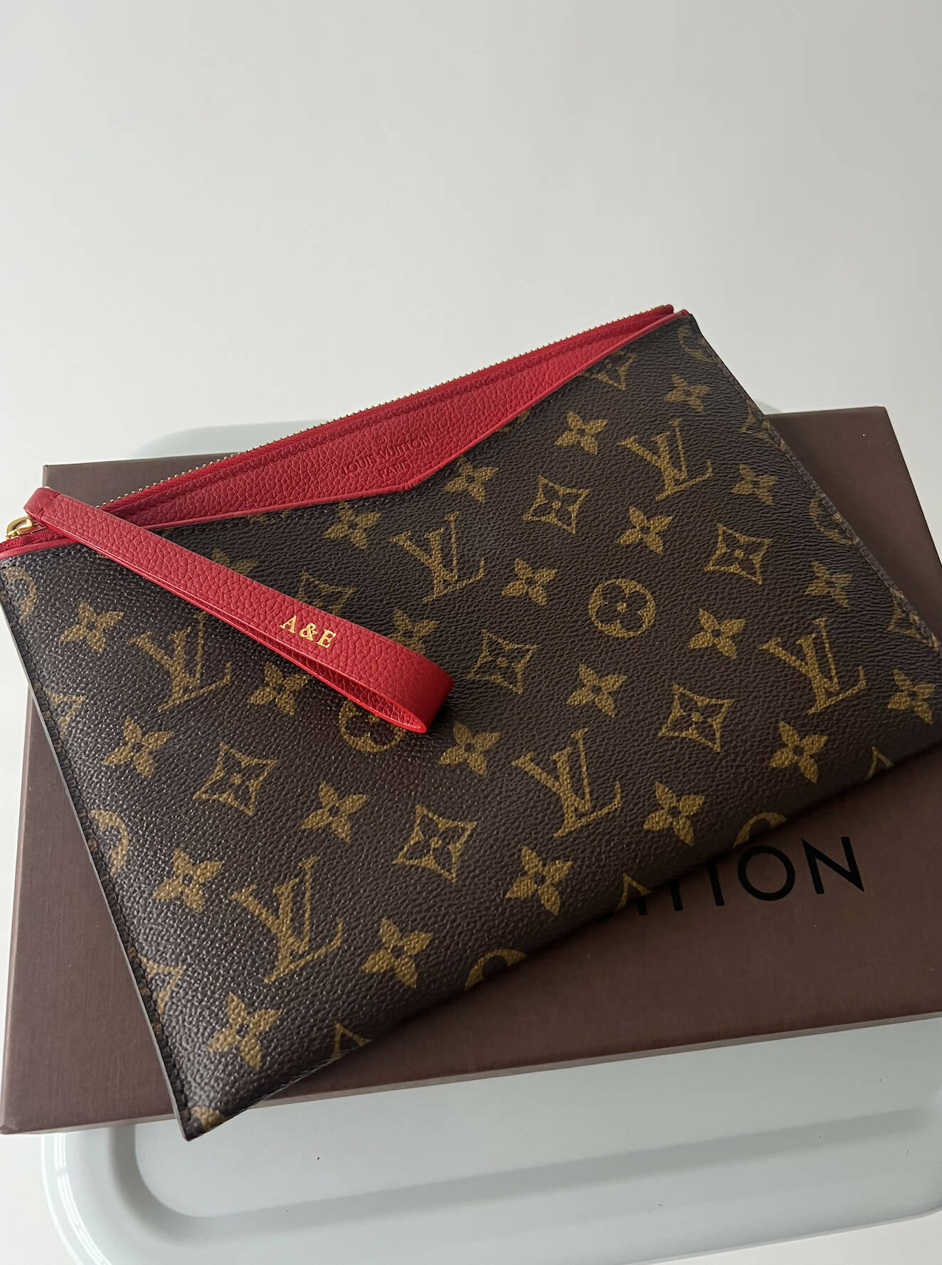 A Louis Vuitton Bicolor Monogram Pallas Pochette by EXCHANGE with a red zipper and wrist strap featuring gold A&E initials rests on a brown box. Receipts included for authenticity.