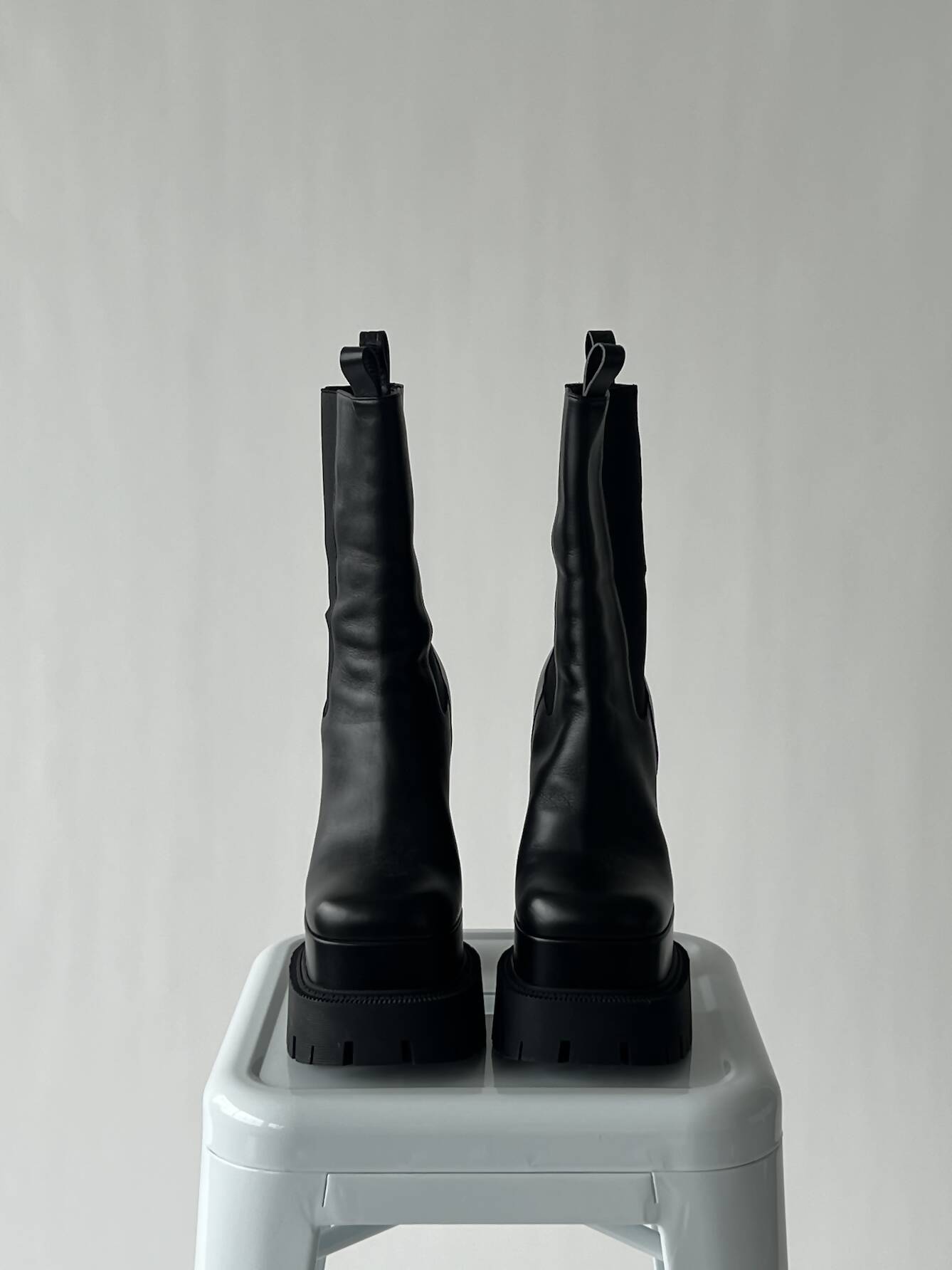 A pair of VERSACE Aevitas Platform Boots - FULL SET by EXCHANGE in size 39, black leather with chunky soles, are placed side by side on a white stool against a plain light gray background.