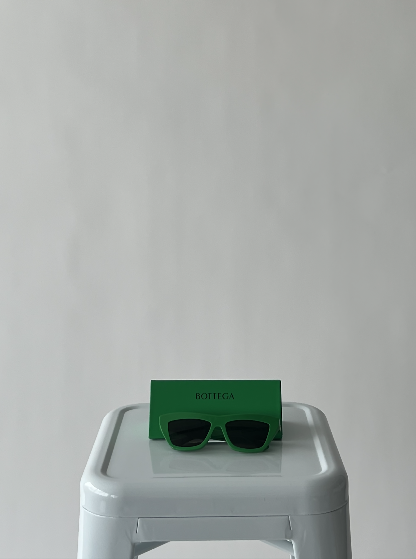 A pair of Bottega Veneta New Entry Cat Eye Sunglasses by EXCHANGE and a matching BOTTEGA case sit on a white stool against a light grey background, exemplifying luxury eyewear.
