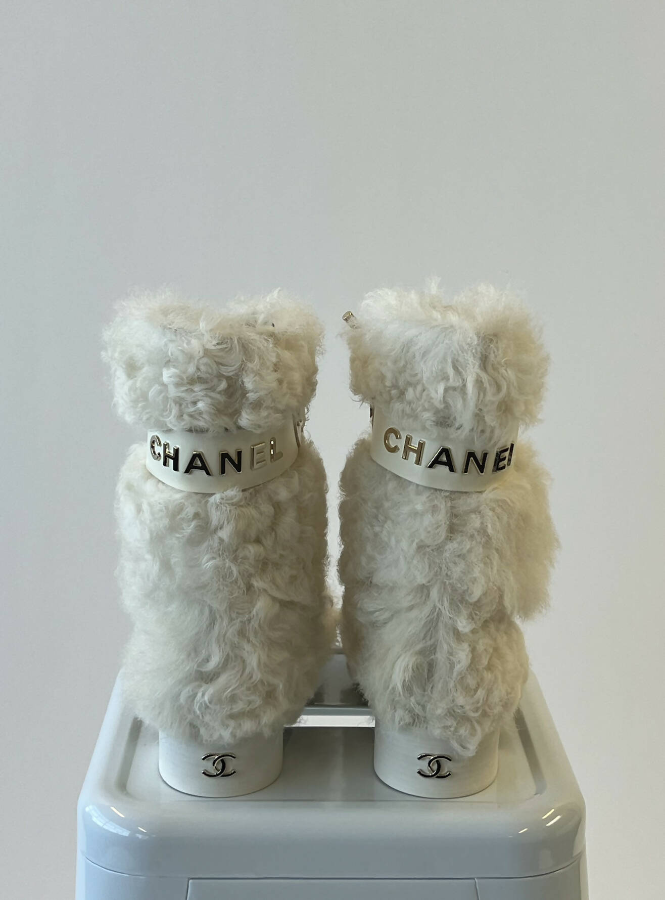 A pair of Chanel White/Black Shearling Fur Calf Length Boots by EXCHANGE, featuring white fluffy fur and gold CHANEL logos on the bands, displayed on a white surface against a plain background.