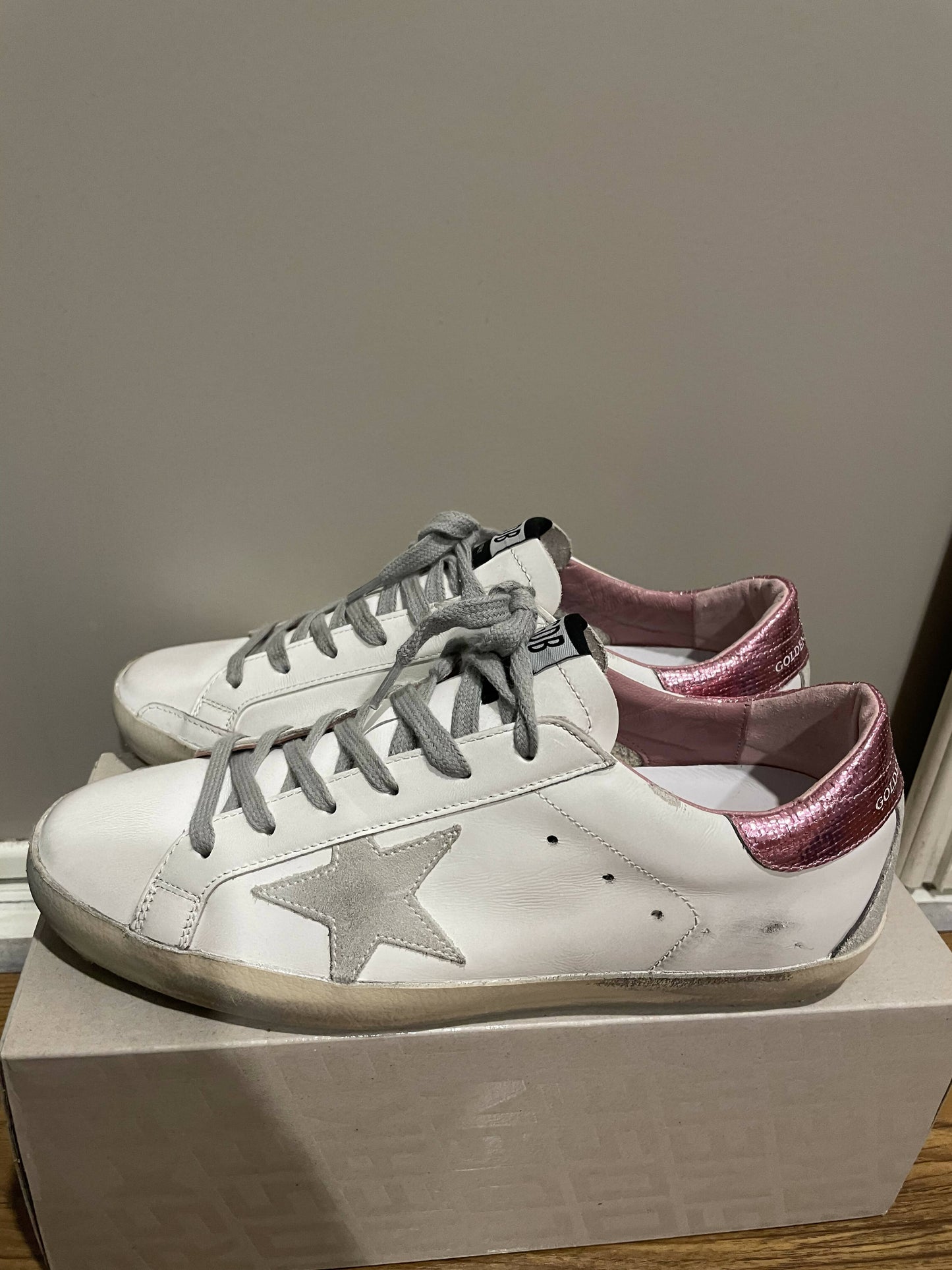 A pair of Golden Goose Superstar sneakers by EXCHANGE, featuring white Italian leather, gray laces, silver star accents, and metallic pink heel tabs, are displayed on a beige shoe box against a light gray wall.