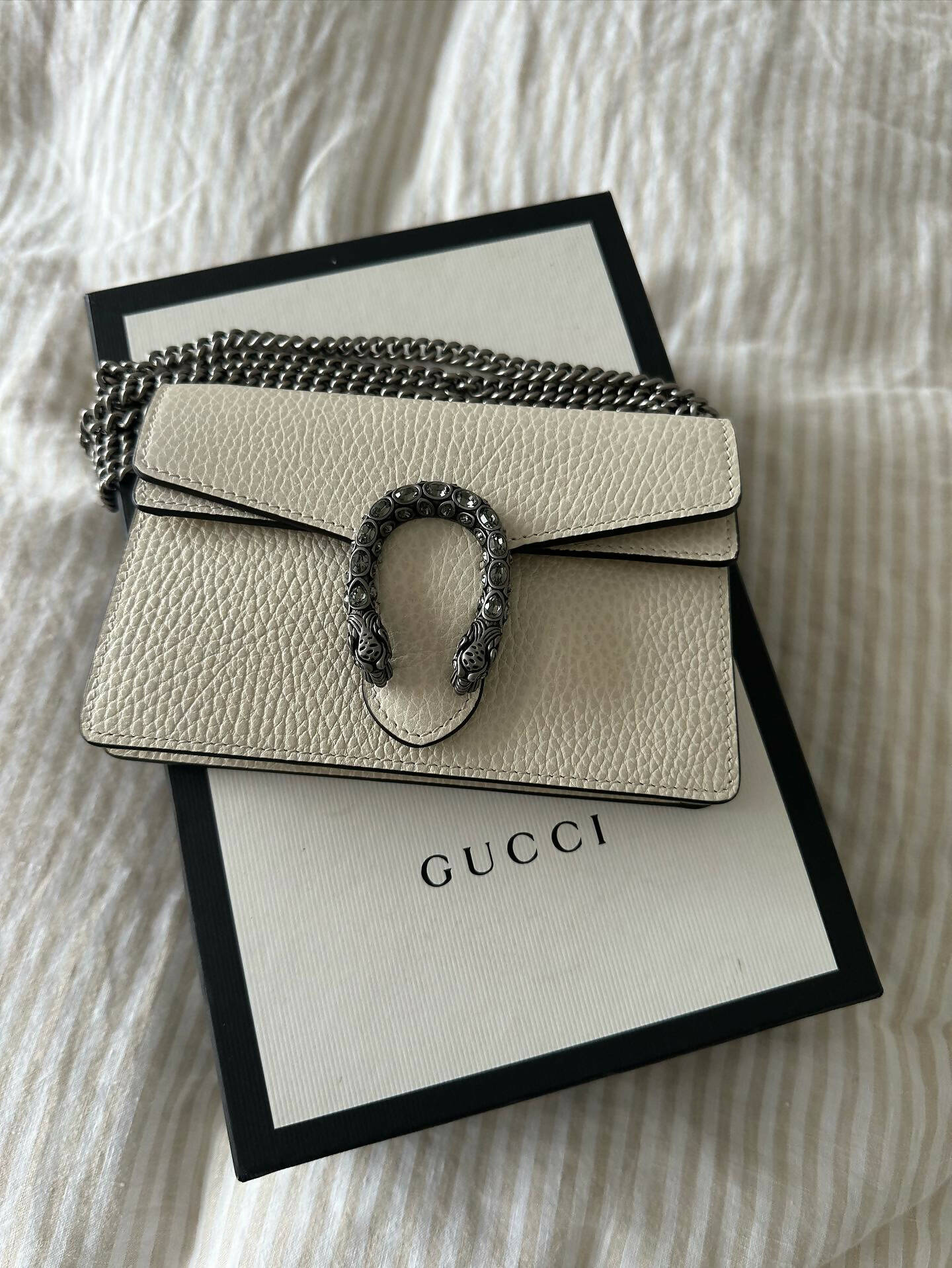 The Gucci Dionysus Super Mini Crossbody Bag - FULL SET by EXCHANGE, featuring genuine white calf leather and a silver chain strap, rests on a white Gucci box with black edges atop a cream fabric background.